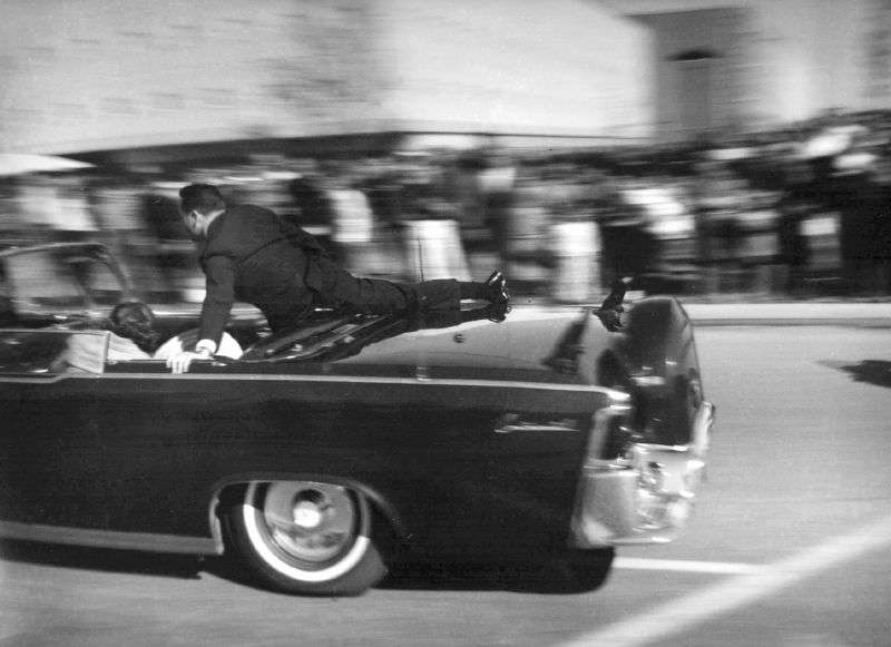 The limousine carrying mortally wounded President John F. Kennedy races toward the hospital seconds after he was shot in Dallas, Nov. 22, 1963. Secret Service agent Clinton Hill is riding on the back of the car.
