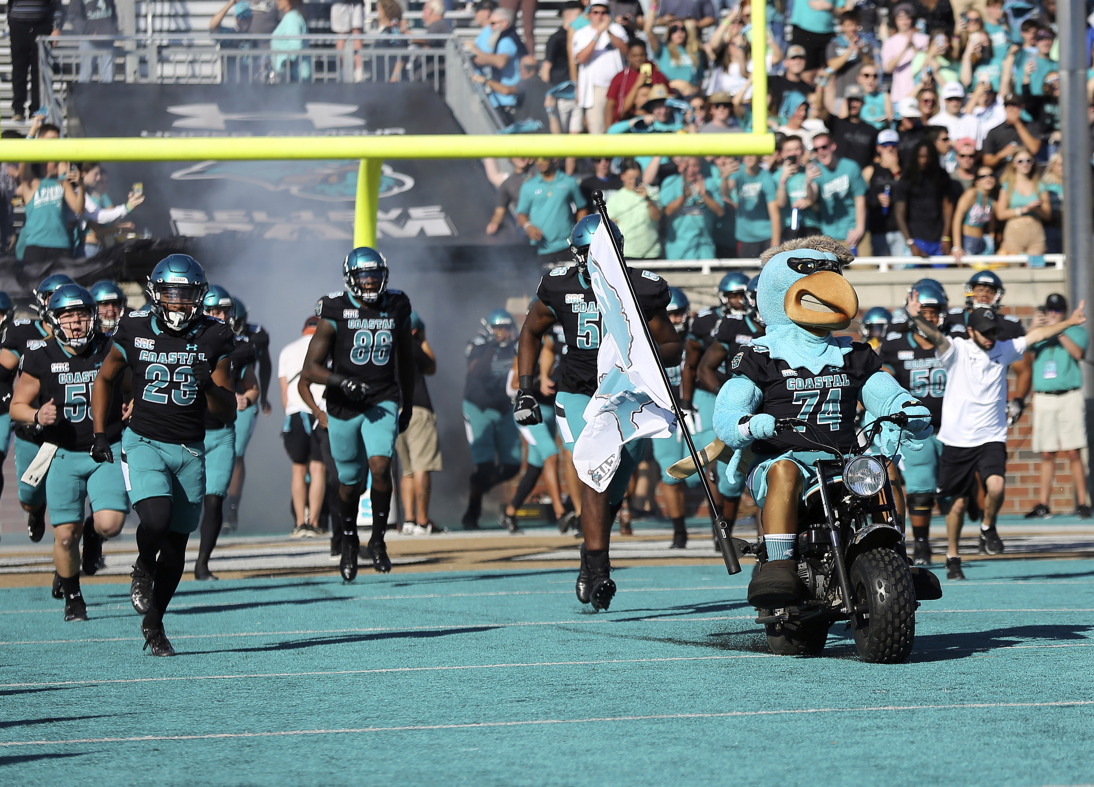 FILE - Mascot Chauncey the Chanticleer, front right, leads Coastal Carolina players onto the field before an NCAA college football game against Georgia State, Nov. 13, 2021, in Conway, S.C. Coastal Carolina's distinctive, “Surf Turf” teal field will feature the logo for the South Carolina State Election Commission this season. 