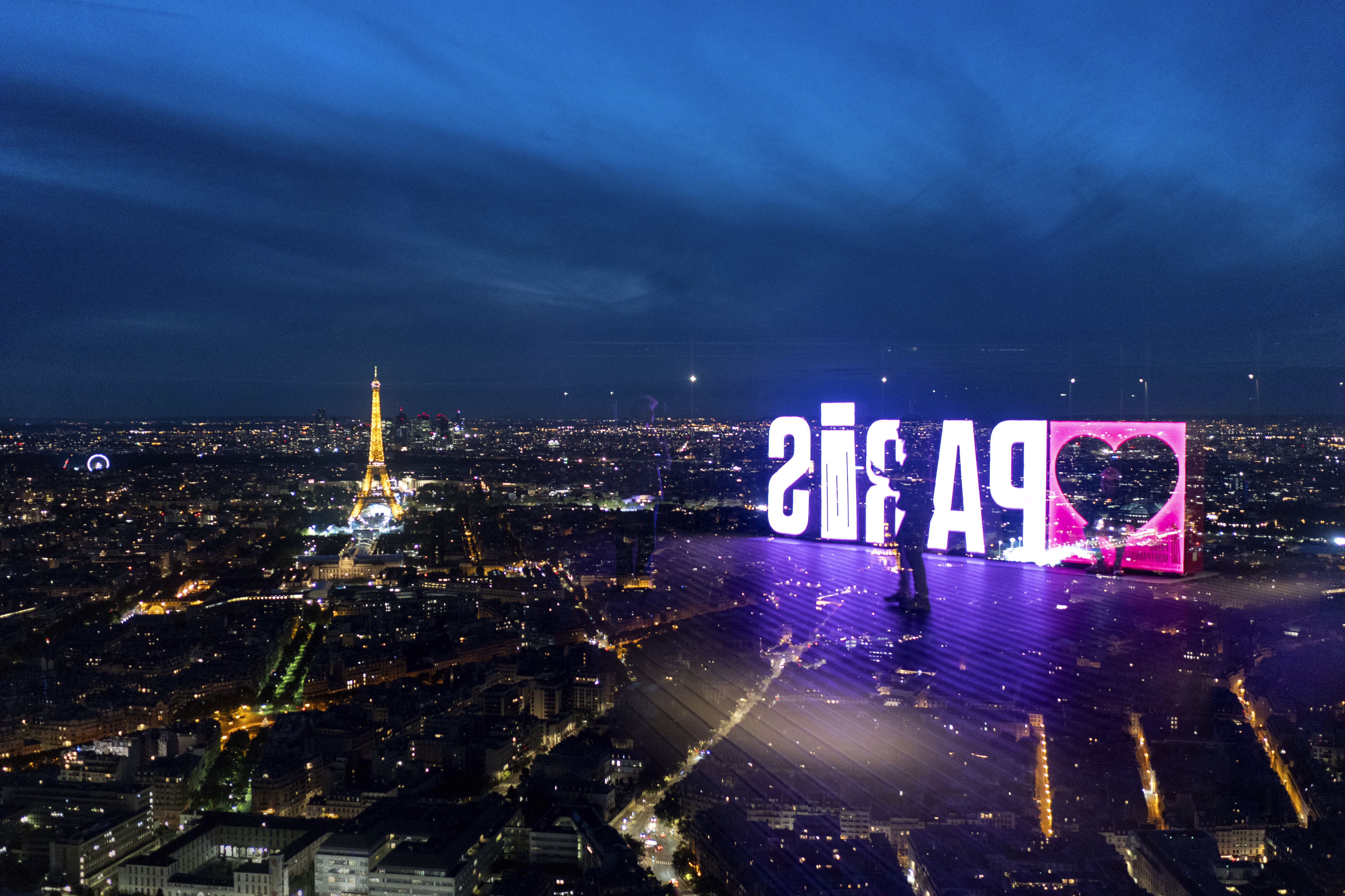 A visitor strolls past a "Paris" sign that is reflected in the glass wall of the Montparnasse tower observation deck as the Eiffel Tower stands in the background ahead of the 2024 Summer Olympics, Tuesday, July 23, 2024, in Paris. 