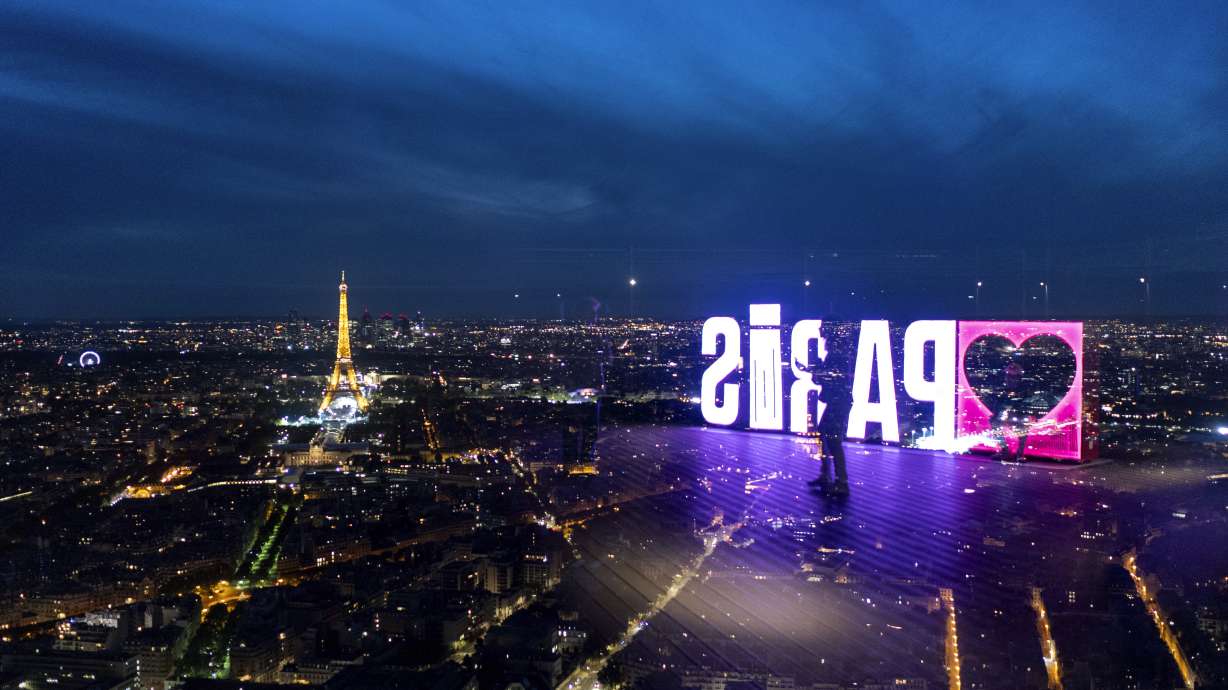 A visitor strolls past a "Paris" sign that is reflected in the glass wall of the Montparnasse tower observation deck as the Eiffel Tower stands in the background ahead of the 2024 Summer Olympics, Tuesday, July 23, 2024, in Paris.