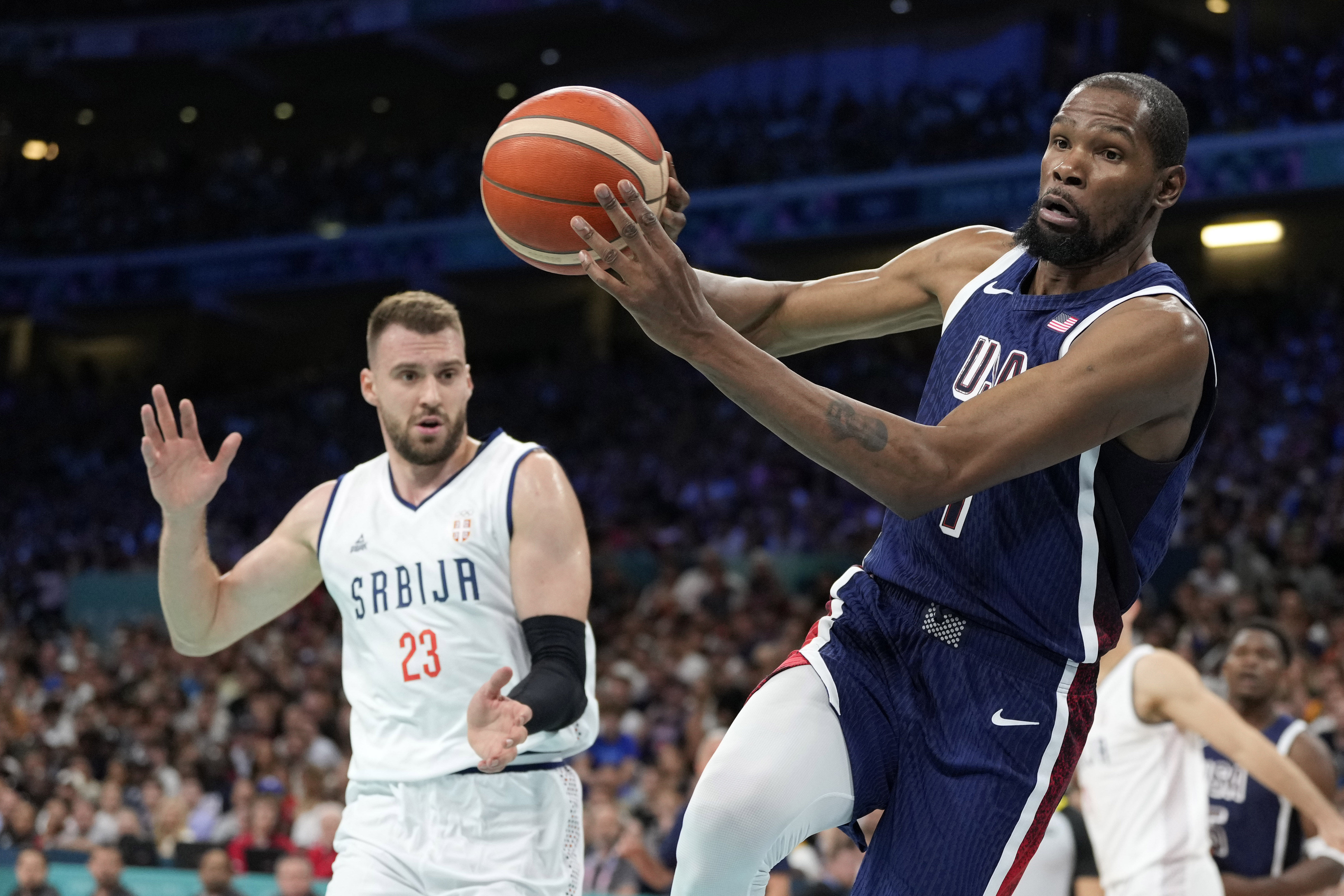 Kevin Durant, right, of the United States, passes the ball as he heads out of bounds while under pressure from Marko Guduric, of Serbia, in a men's basketball game at the 2024 Summer Olympics, Sunday, July 28, 2024, in Villeneuve-d'Ascq, France. 