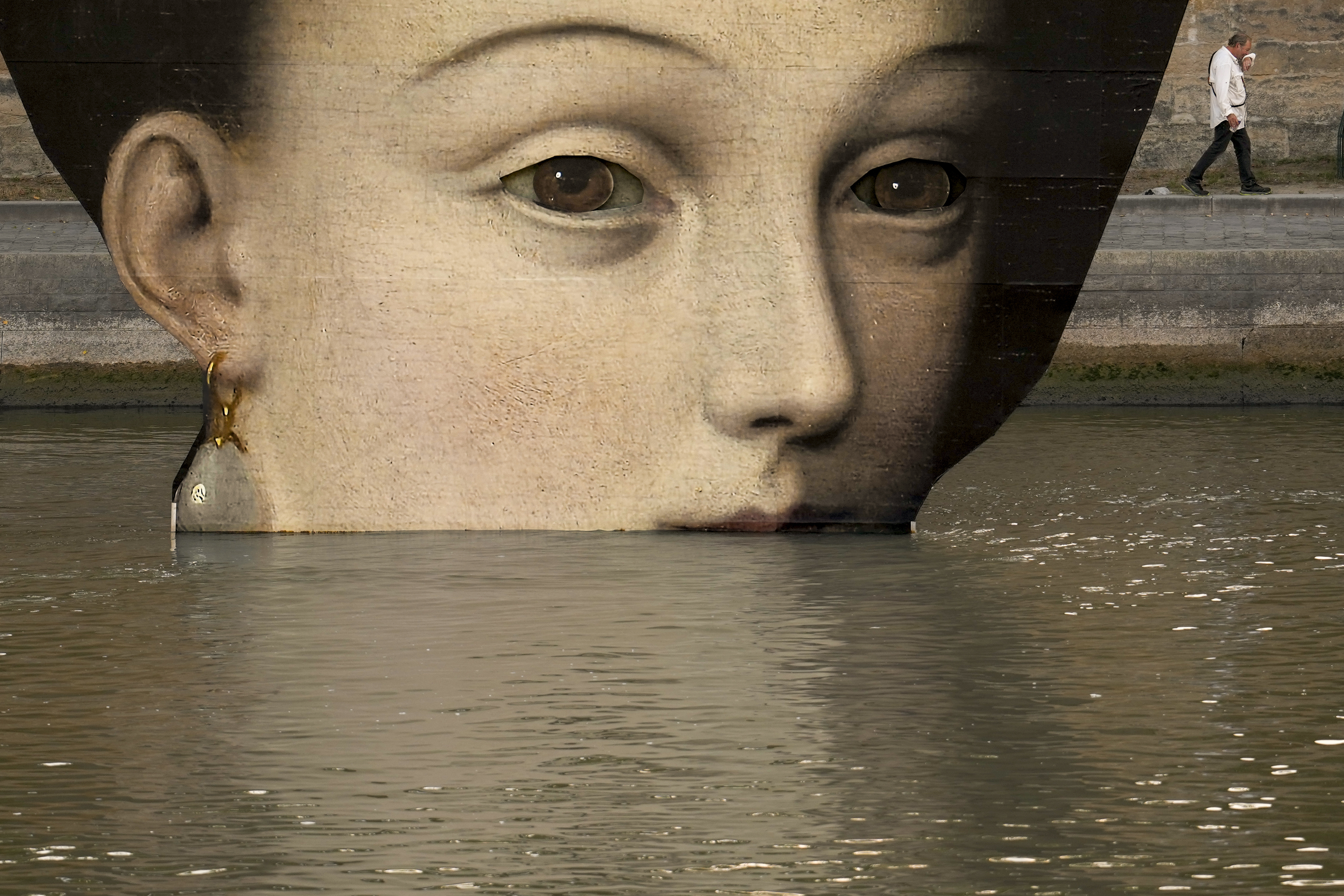 A man wipes his face as he walks past reproductions of artworks decorating the banks of the River Seineat the 2024 Summer Olympics, Tuesday, July 30, 2024, in Paris, France. The men's Olympic triathlon has been postponed over concerns about water quality in Paris' Seine River, where the swimming portion of the race was supposed to take place. 