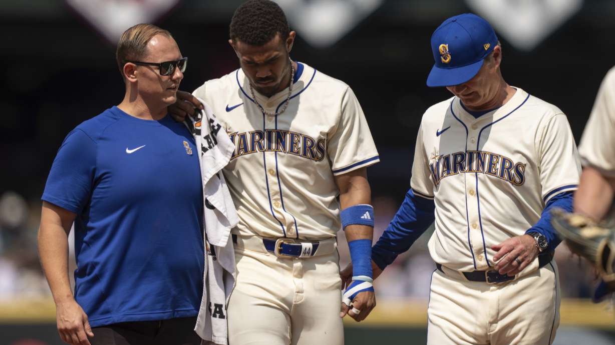 El jardinero central dominicano de los Marineros de Seattle, Julio Rodríguez (centro) es ayudado a salir del campo por el mánager Scott Servais (derecha), y por un integrante del cuerpo técnico durante la sexta entrada del juego de béisbol ante los Astros de Houston, el domingo 21 de julio de 2024, en Seattle.