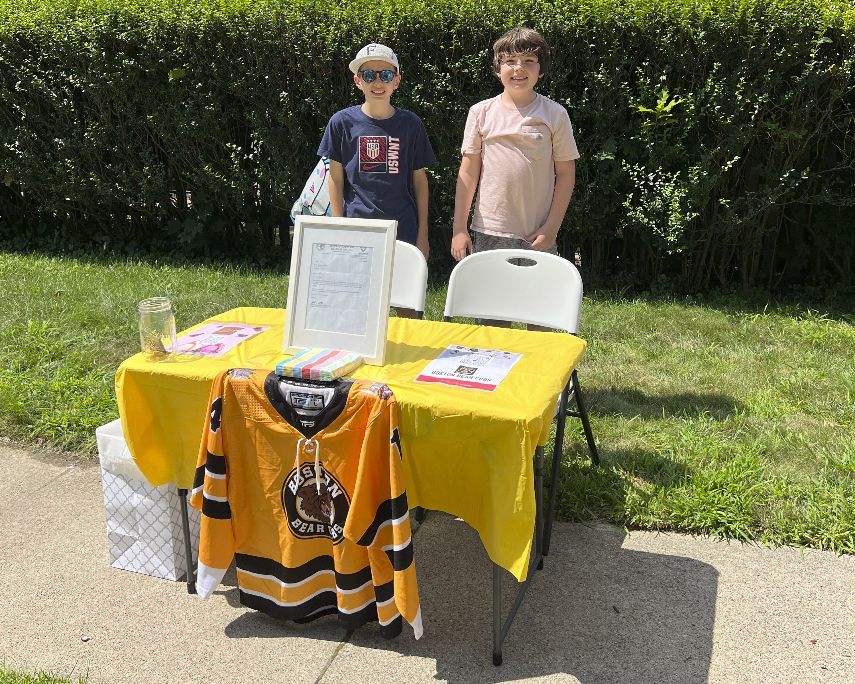In this Saturday, Aug. 10, 2024 photo provided by Meghan Doherty, Ben Doherty, 12, of Braintree, Mass., left, stands with his cousin Danny Doherty, 12, of Norwood, Mass., right, near a table for donations to the Boston Bear Cubs hockey team, in Norwood.