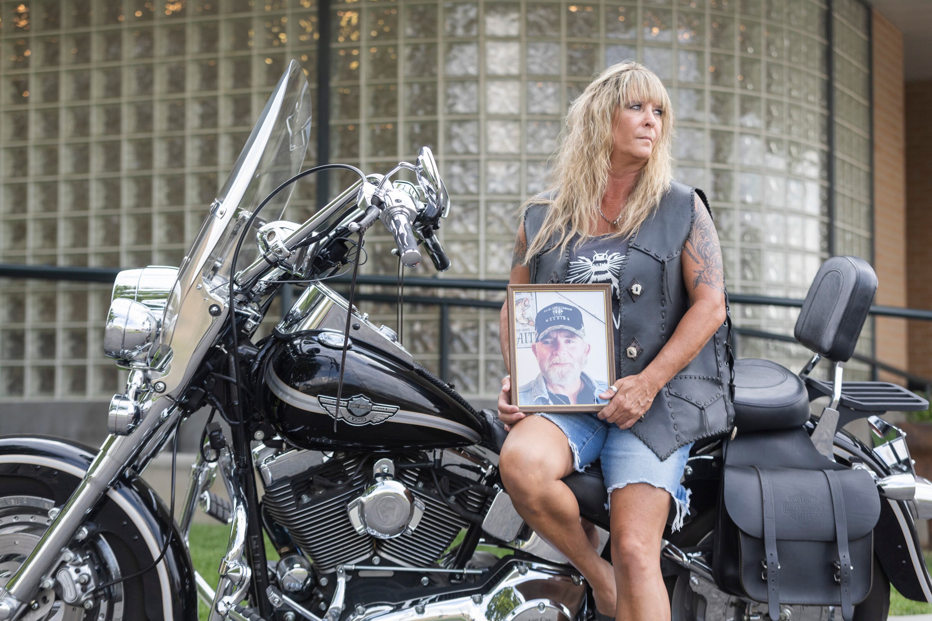 Yvette Shields poses with a photo of her late husband, Larry Shields, on his Harley-Davidson Fat Boy Softail motorcycle while wearing his custom leatherworked riding jacket in front of Tate Mortuary in Tooele on Tuesday.