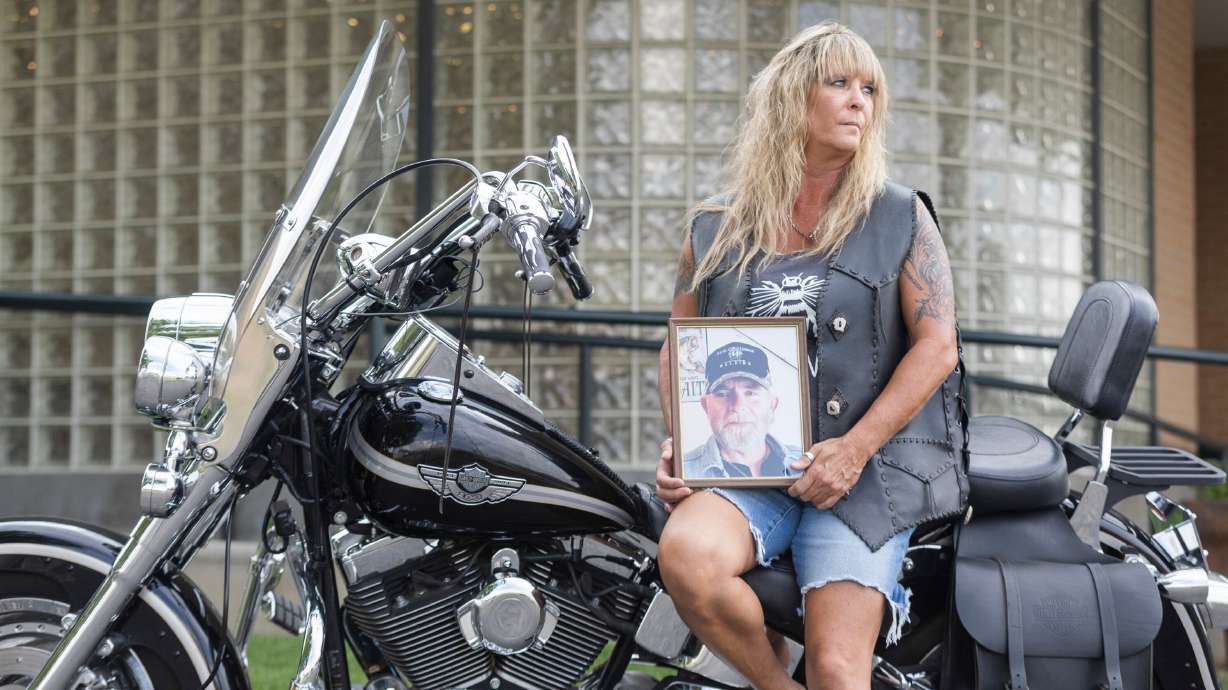 Yvette Shields poses with a photo of her late husband, Larry Shields, on his Harley-Davidson Fat Boy Softail motorcycle while wearing his custom leatherworked riding jacket in front of Tate Mortuary in Tooele on Tuesday.