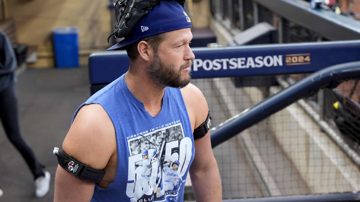 Los Angeles Dodgers pitcher Clayton Kershaw wears a glove on his head and devices on his arms in the dugout during practice Monday, Oct. 7, 2024, a day before Game 3 of a baseball NL Division Series against the San Diego Padres in San Diego.