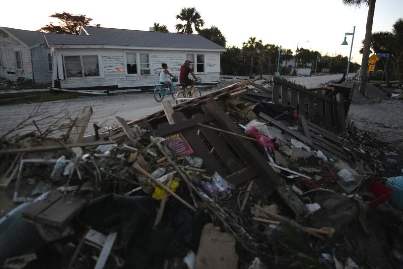 People bike past damaged homes and debris left by Hurricane Milton, on the sand-coated main road of southern Manasota Key, already cleared of feet of sand, in Englewood, Fla., Sunday.