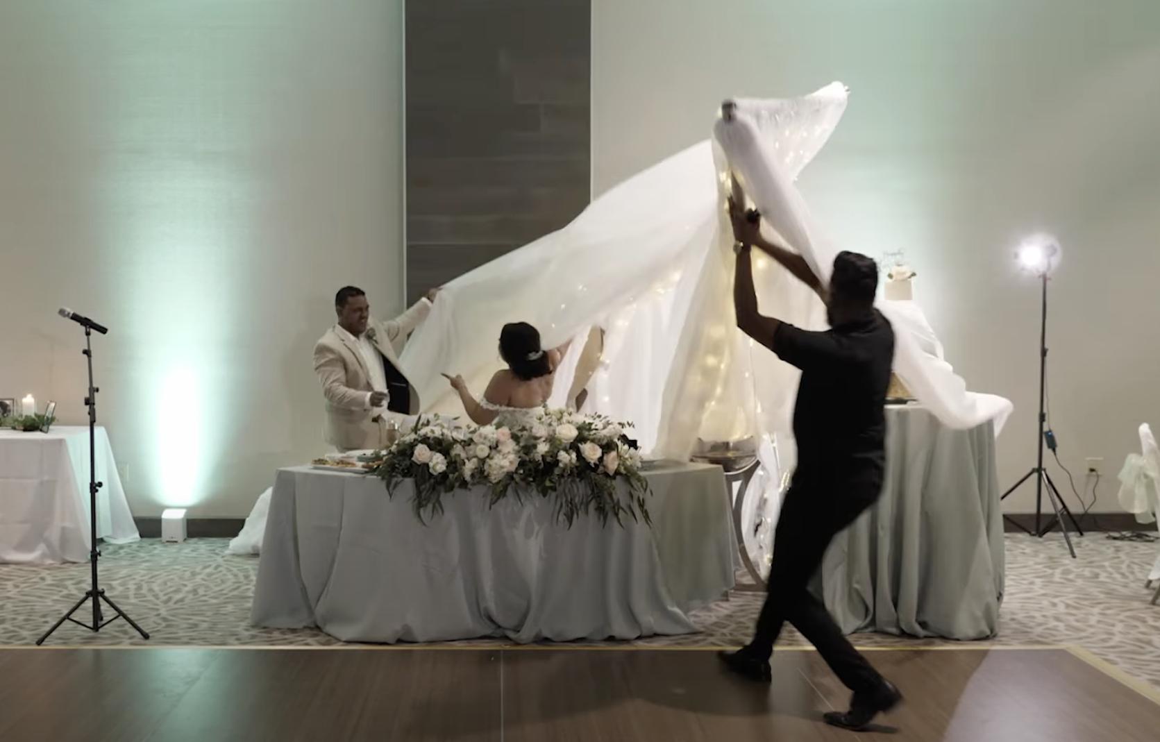 A man finishes a best man speech at a wedding, and as he hugs the bride and groom, the backdrop falls on them and the cake.