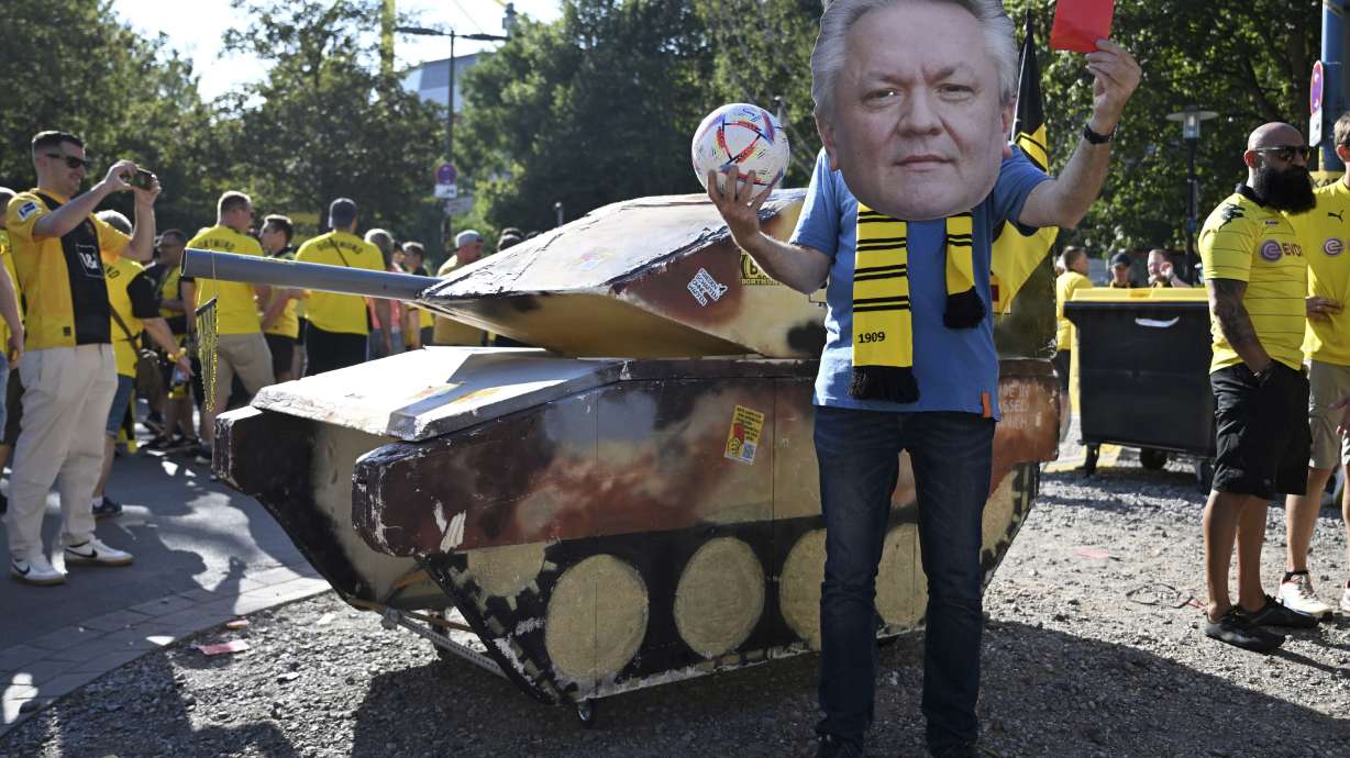FILE - A dummy tank stands next to an activist wearing a mask with the likeness of Rheinmetall CEO Armin Papperger and holding up a red card ahead of the soccer game between Borussia Dortmund and Eintracht Frankfurt, in Dortmund, Germany, Saturday Aug. 24, 2024.