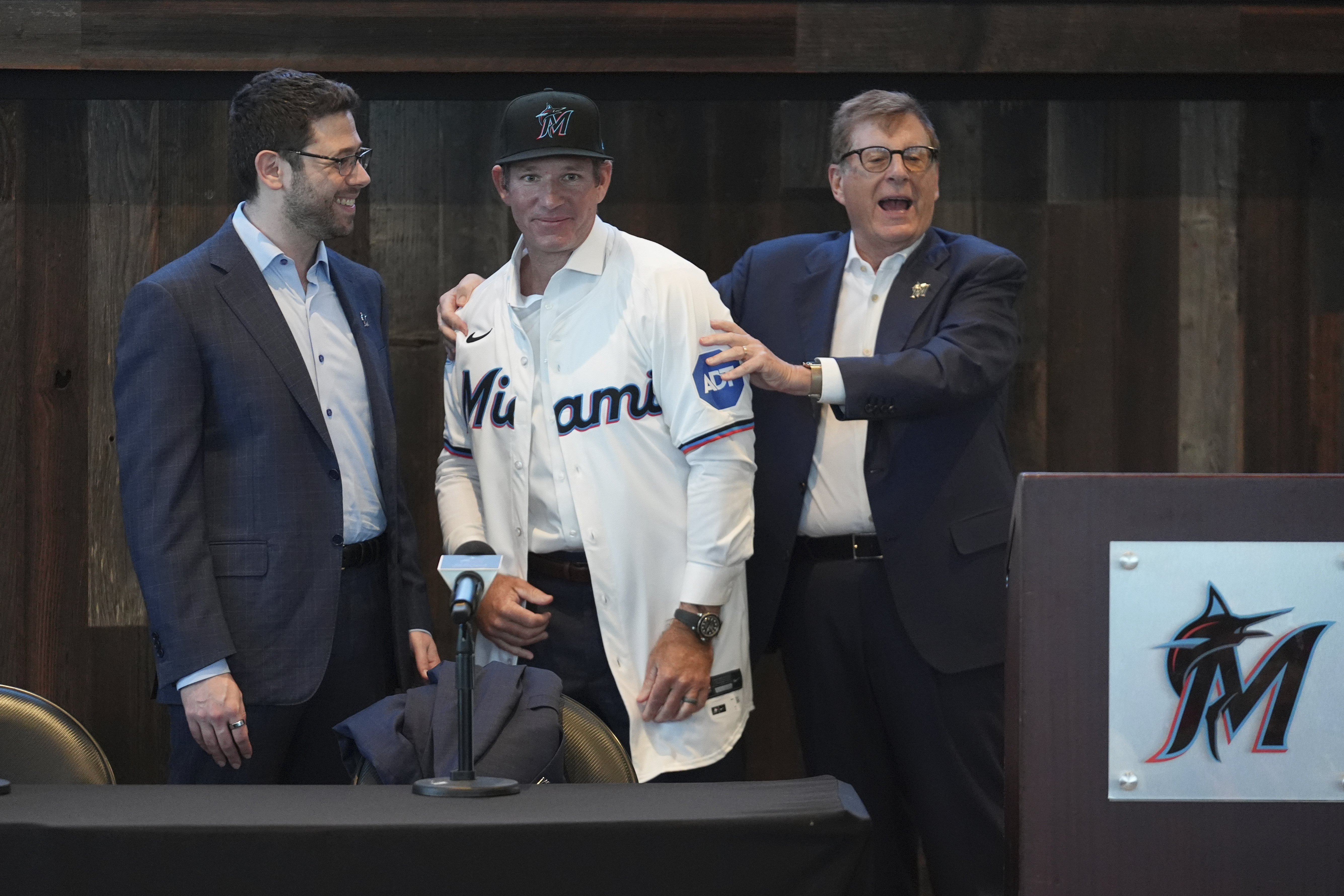Clayton McCullough, center, puts on a Miami Marlins jersey and cap as he is presented by Marlins owner Bruce Sherman, right, and Peter Bendix, president of baseball operations, Monday, Nov. 18, 2024, in Miami.