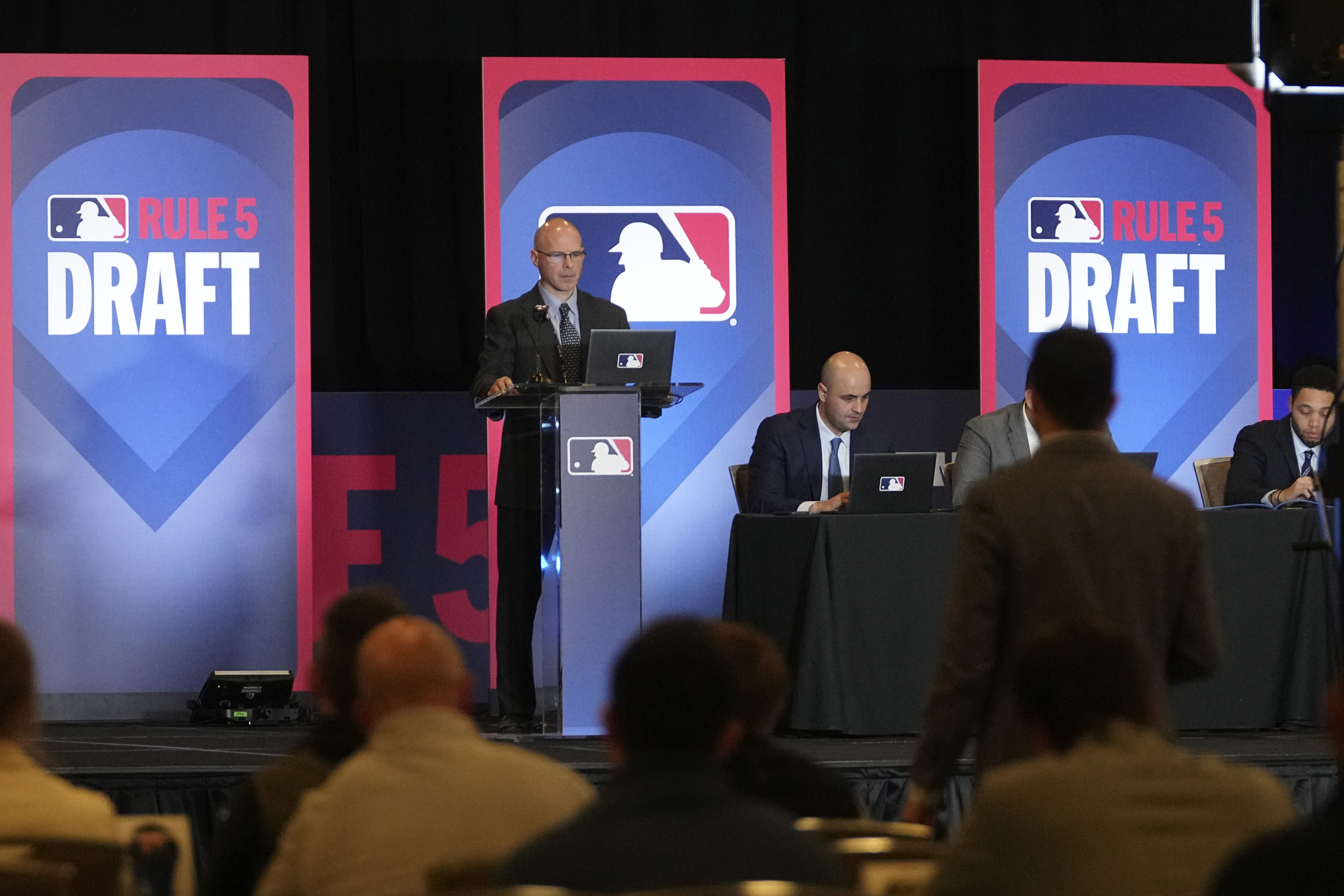 Jeff Pfeifer, at podium, MLB vice president of baseball operations, listens to a pick made by a San Diego Padres representative, standing right, during the Rule 5 Draft at the Major League Baseball winter meetings in Dallas, Wednesday, Dec. 11, 2024.