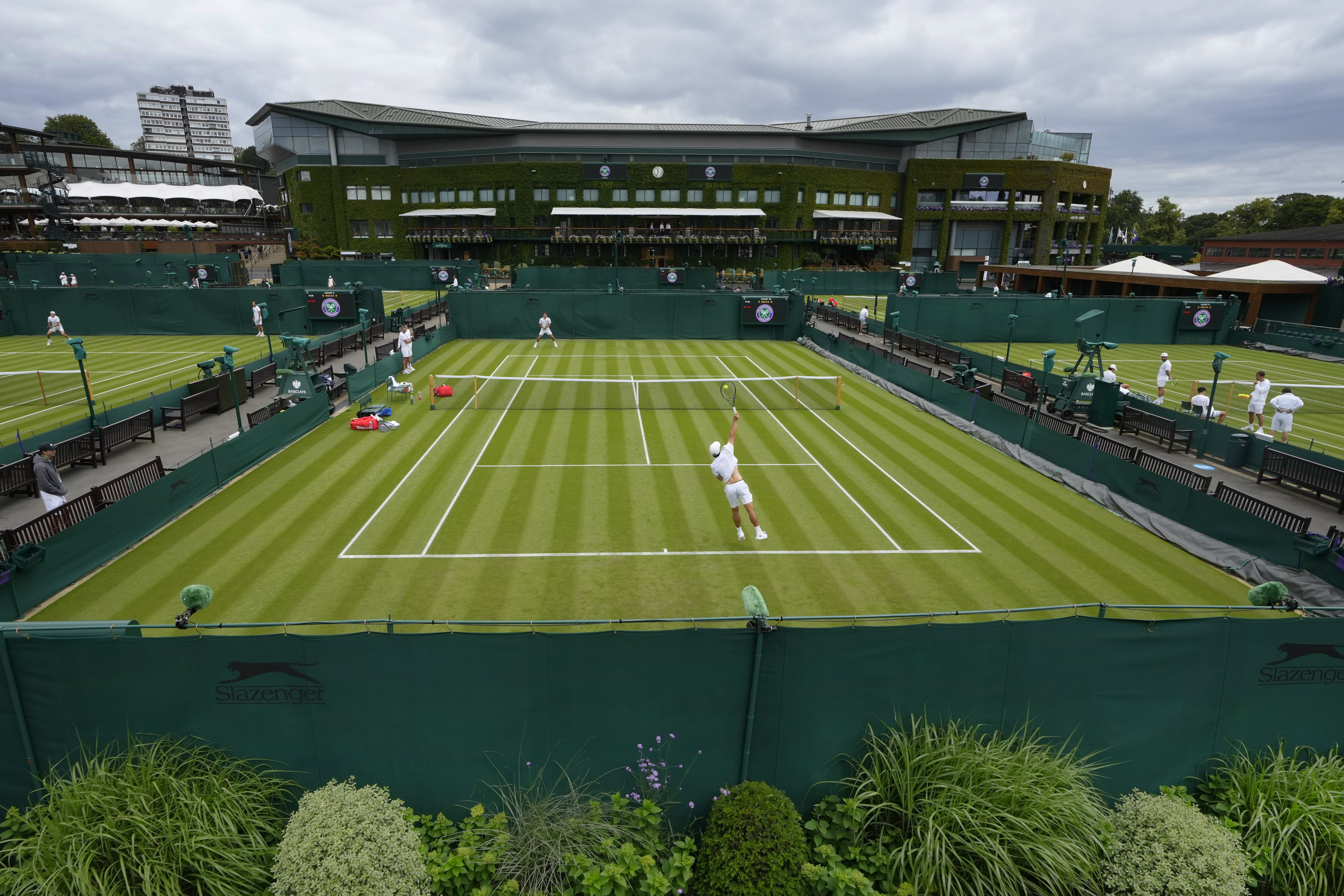 FILE - Tennis players train as a member of ground staff waters the plants at the All England Lawn Tennis and Croquet Club in Wimbledon, London, Friday, June 28, 2024.