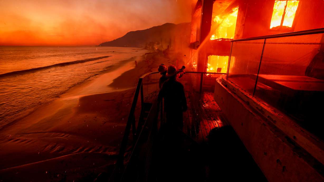Firefighters work from a deck as the Palisades Fire burns a beachfront property, Wednesday, in Malibu, Calif.
