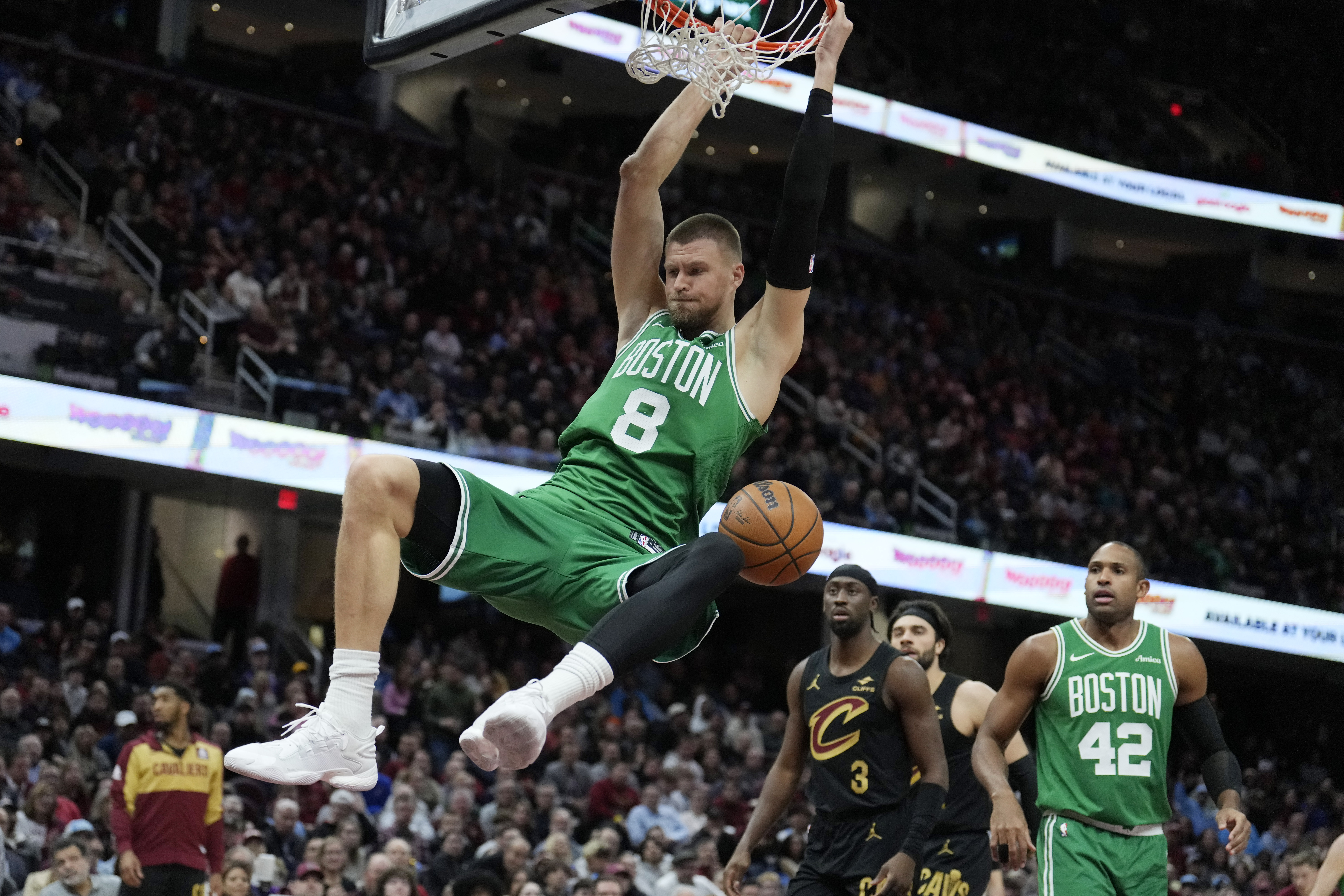 Boston Celtics center Kristaps Porzingis (8) hangs from the basket in front of Cleveland Cavaliers guard Caris LeVert (3) after a dunk in the second half of an NBA basketball game, Tuesday, Feb. 4, 2025, in Cleveland.