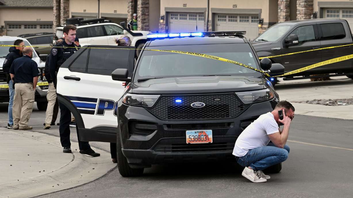 An unidentified man crouches down in front of a police vehicle talking on a cellphone as Saratoga Springs police investigate the death of an 8-year-old boy and the injury of a woman in a home on Friday.
