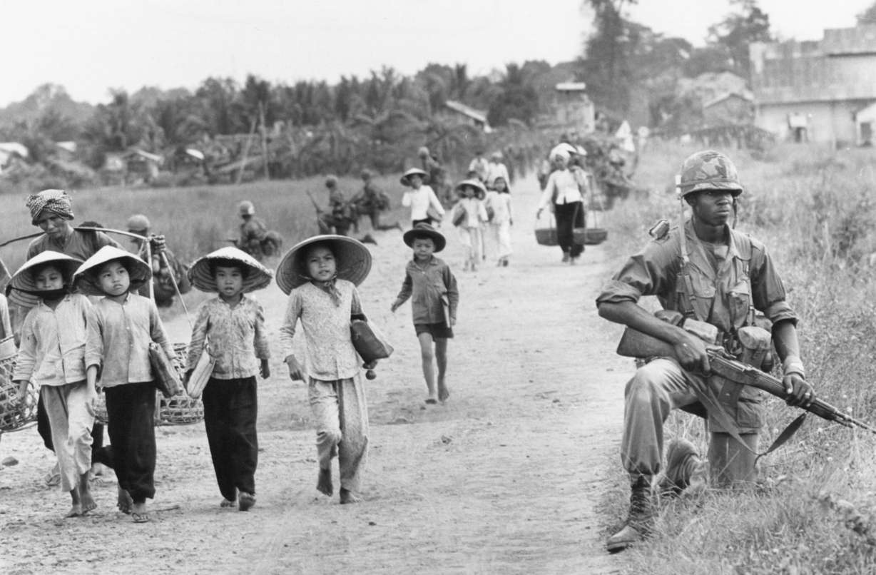 In an area heavily infiltrated by Viet Cong, a U.S. 1st Division soldier guards Route 7 as Vietnamese market women and schoolchildren return home to the village of Xuan Dien from Ben Cat, December 1965.