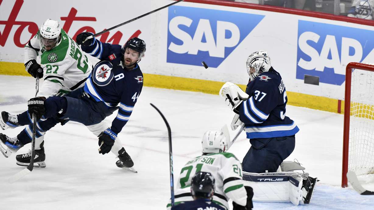 Winnipeg Jets goaltender Connor Hellebuyck (37) makes a save as Dallas Stars' Mason Marchment (27) dumps Jets' Josh Morrissey (44) during the first period of Game 2 of a second-round NHL hockey playoff series in Winnipeg, Manitoba, Friday, May 9, 2025.