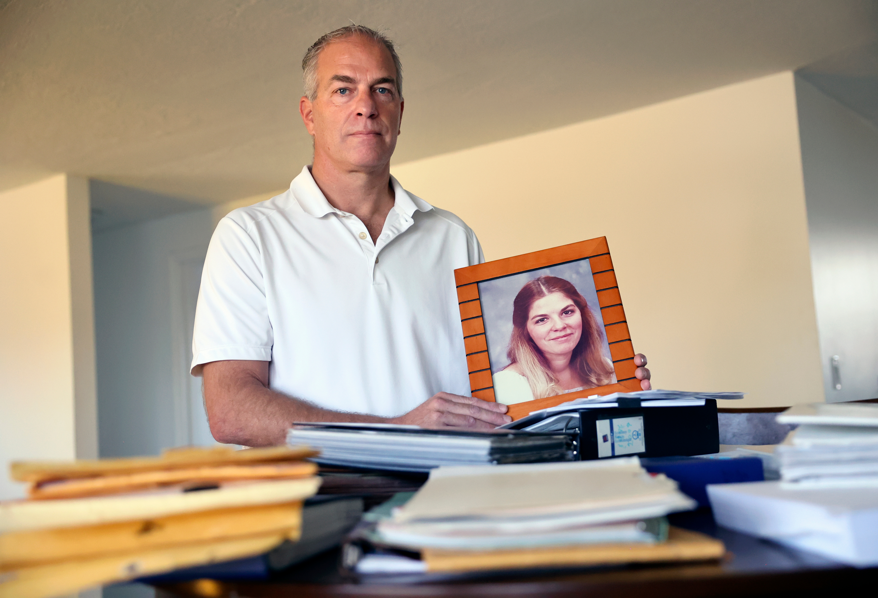 Matt Hunsaker poses for a portrait with a photo of his mother, Maurine Hunsaker, who was murdered in 1986, and stacks of paperwork related to the case, at his home in Taylorsville on Jan. 30, 2024.
