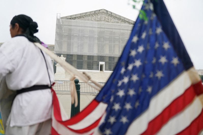 A person stands with a U.S. flag attached to them outside the Supreme Court in Washington, June 27. The court will once again decide the legality of President Donald Trump's birthright citizenship order, it announced on Friday.