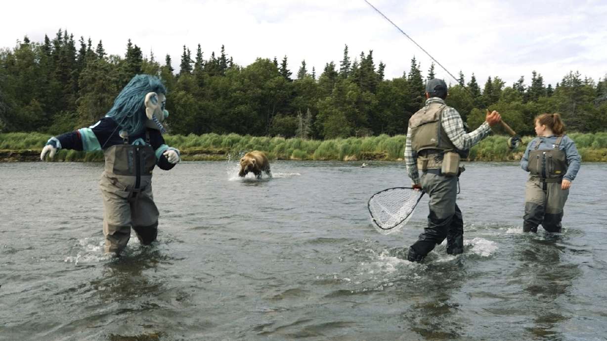 This image taken from video provided by the Seattle Kraken shows a brown bear splashing toward the NHL Kraken sea troll mascot known as Buoy, left, next to a fishing guide during the filming of a promotional video in a river outside Anchorage, Alaska, June 25, 2025.