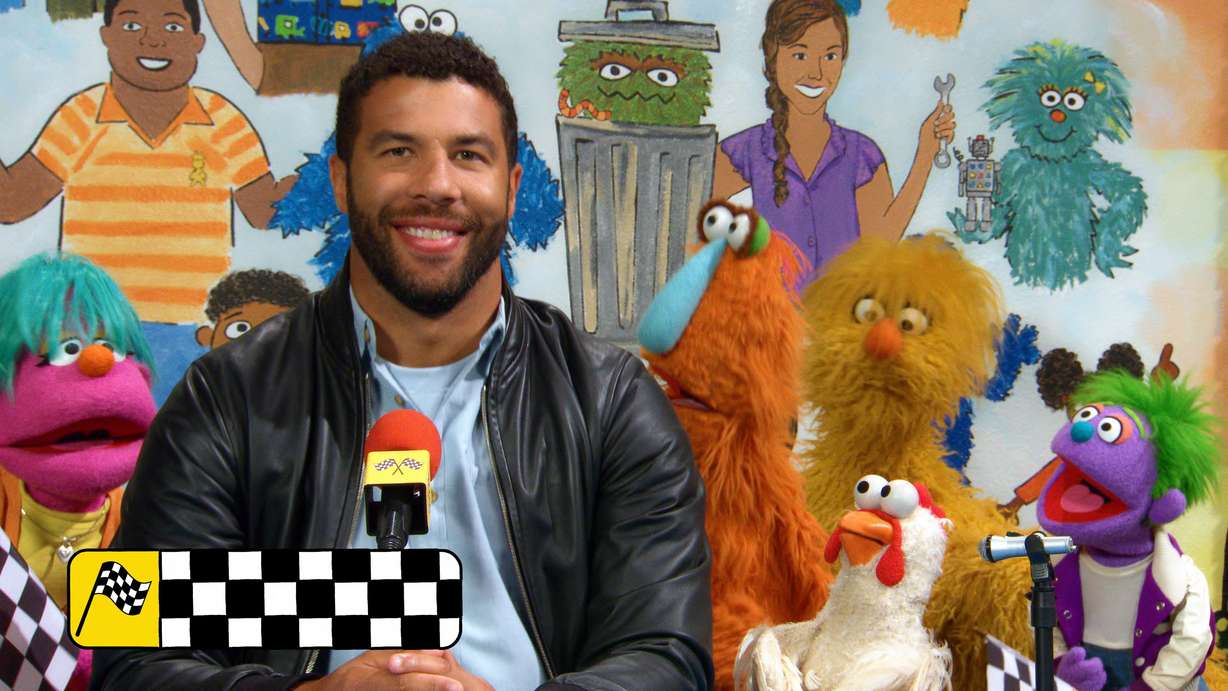 This image shows Bubba Wallace with various muppet characters on the set of "Sesame Street."