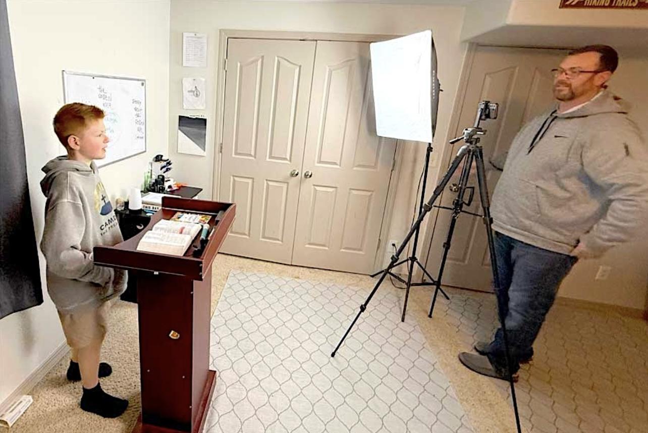 Camden Pruett, left, has his father, Greg Pruett, record a sermon on “The Golden Rule” on Sunday from his bedroom/studio in his home in Rigby.