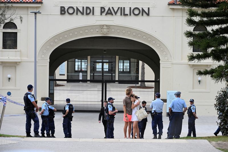 Two people embrace as police officers stand guard outside Bondi Pavilion following the attack on a Jewish holiday celebration at Bondi Beach, in Sydney, Australia, Sunday. A court filing on Wednesday named Naveed Akram as charged with 59 offenses, including 15 murders and terrorism.