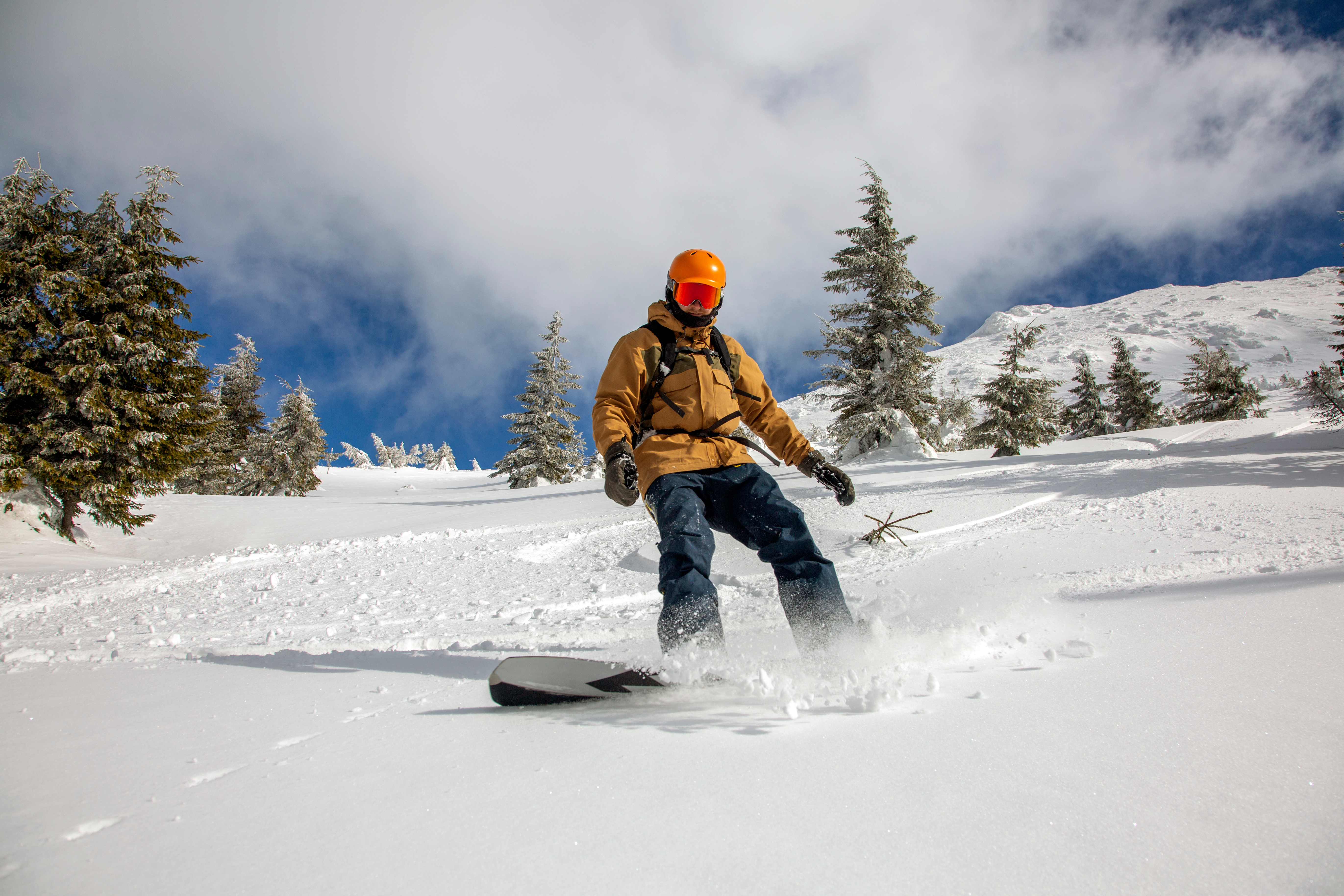 Young athletic snowboarder in bright yellow coat riding down on splitboard splashing snow. Skitouring freeride concept