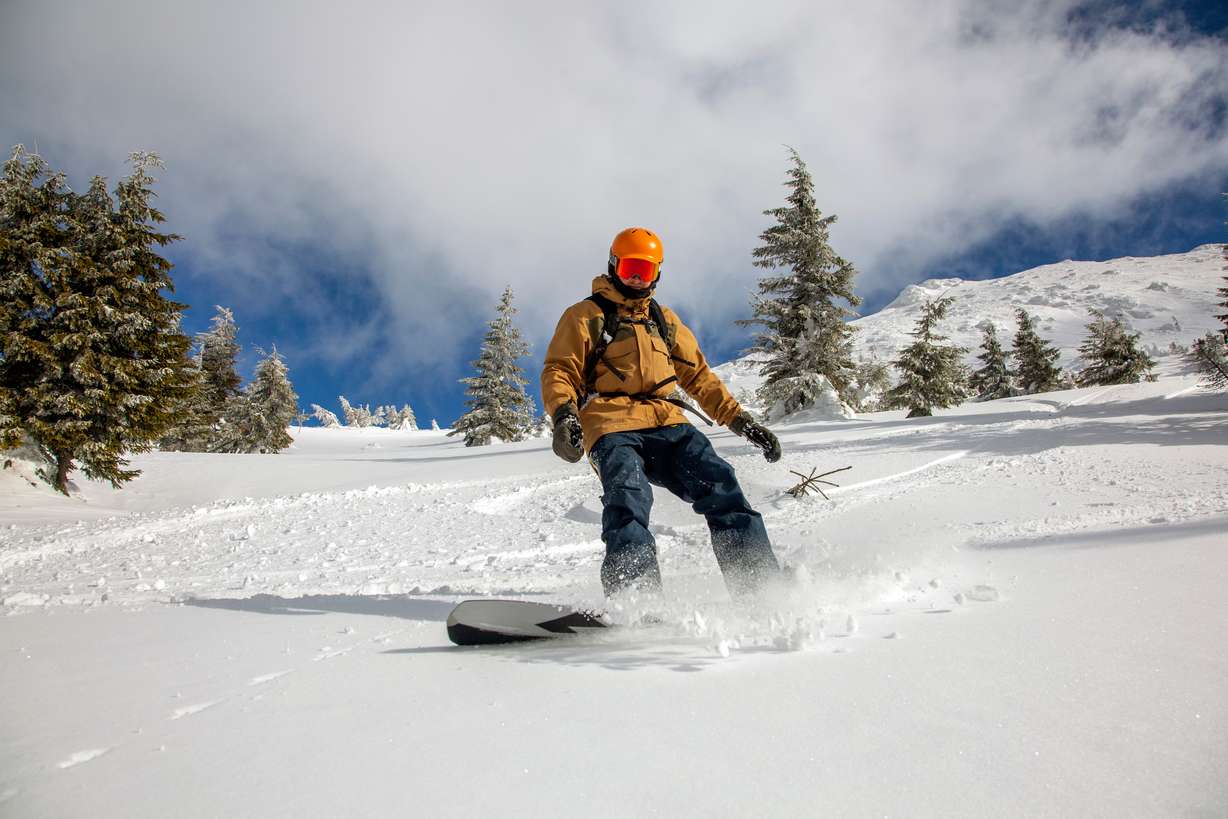 Young athletic snowboarder in bright yellow coat riding down on splitboard splashing snow. Skitouring freeride concept