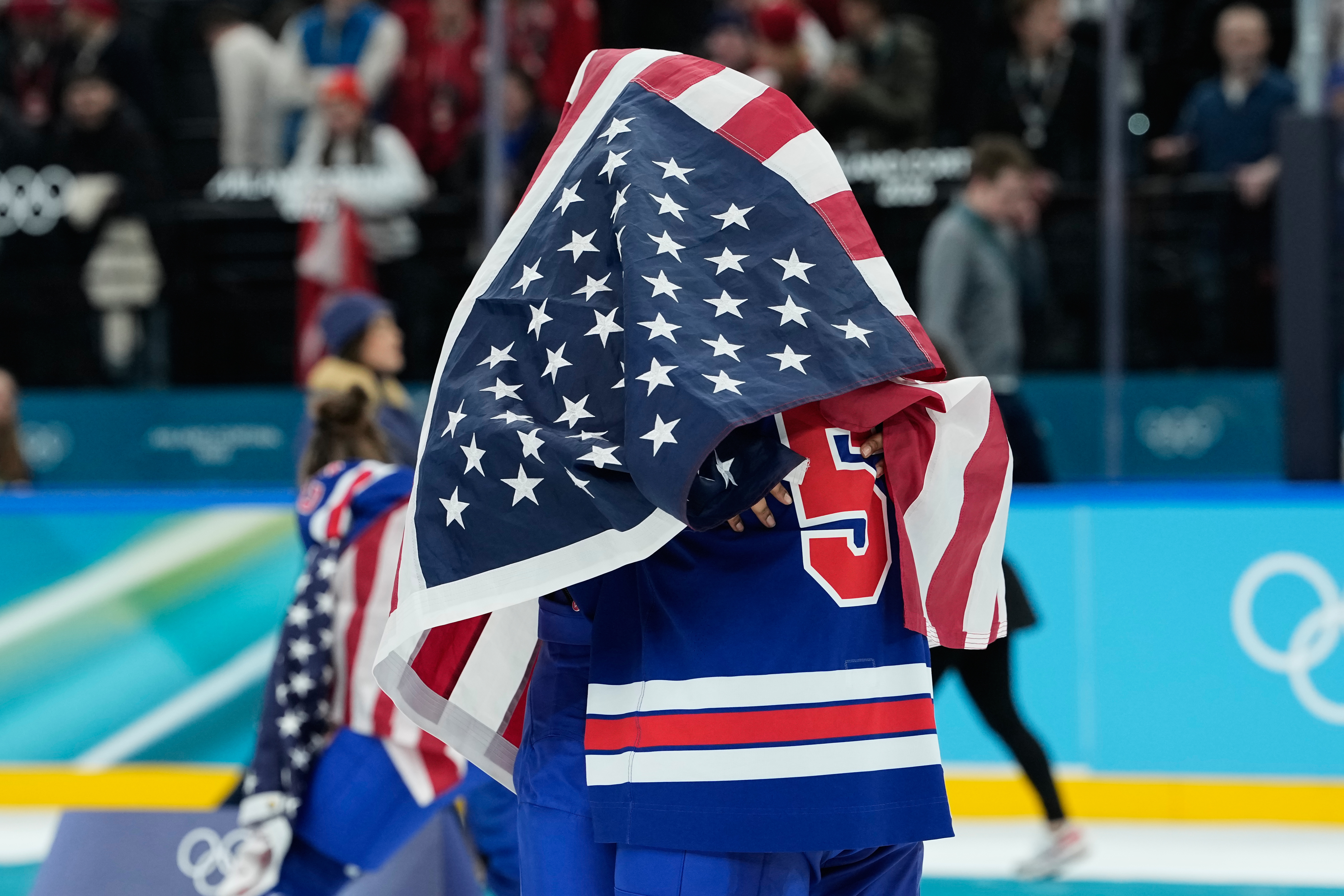 United States' Megan Keller (5), covered in the U.S. flag, gets a hug from a teammate after the United States' women's ice hockey team stand after being presented with the gold medals at the 2026 Winter Olympics, in Milan, Italy, Thursday, Feb. 19, 2026.