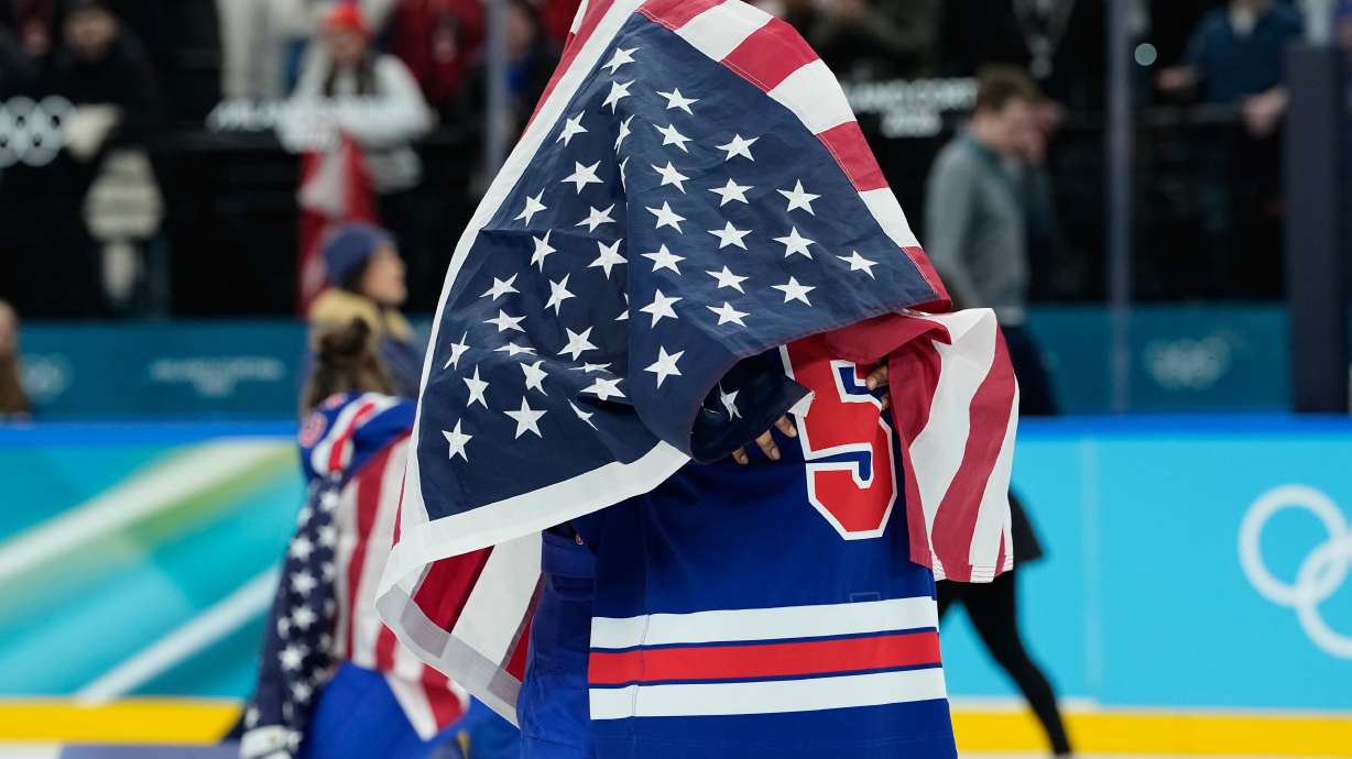 United States' Megan Keller (5), covered in the U.S. flag, gets a hug from a teammate after the United States' women's ice hockey team stand after being presented with the gold medals at the 2026 Winter Olympics, in Milan, Italy, Thursday, Feb. 19, 2026.