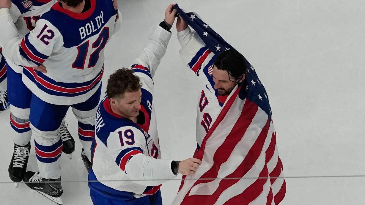 United States' Matthew Tkachuk (19) helps put an American flag around goalie Connor Hellebuyck after the United States beat Canada in overtime to win the men's gold medal hockey game at the 2026 Winter Olympics, in Milan, Italy, Sunday, Feb. 22, 2026.