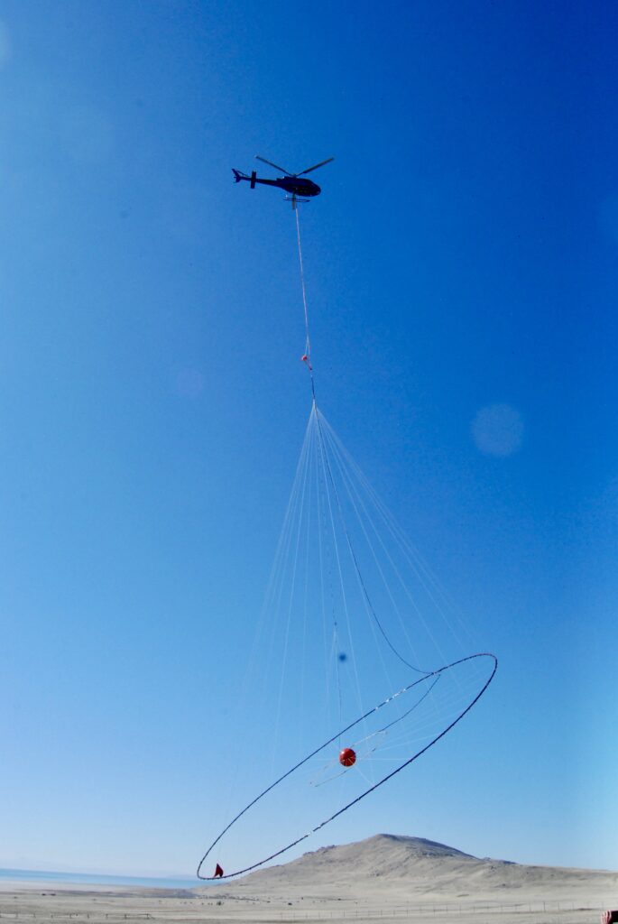 A helicopter lifts off from Antelope Island carrying electromagnetic survey equipment for a geophysical data-gathering mission over Farmington Bay in February 2025.