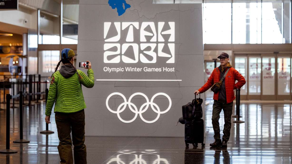 Virag White takes a photo of her husband, Michael, at the “Utah 2034” Olympics logo at the Salt Lake City International Airport on Feb. 20. A U.S. bank could become a new worldwide Olympic sponsor.
