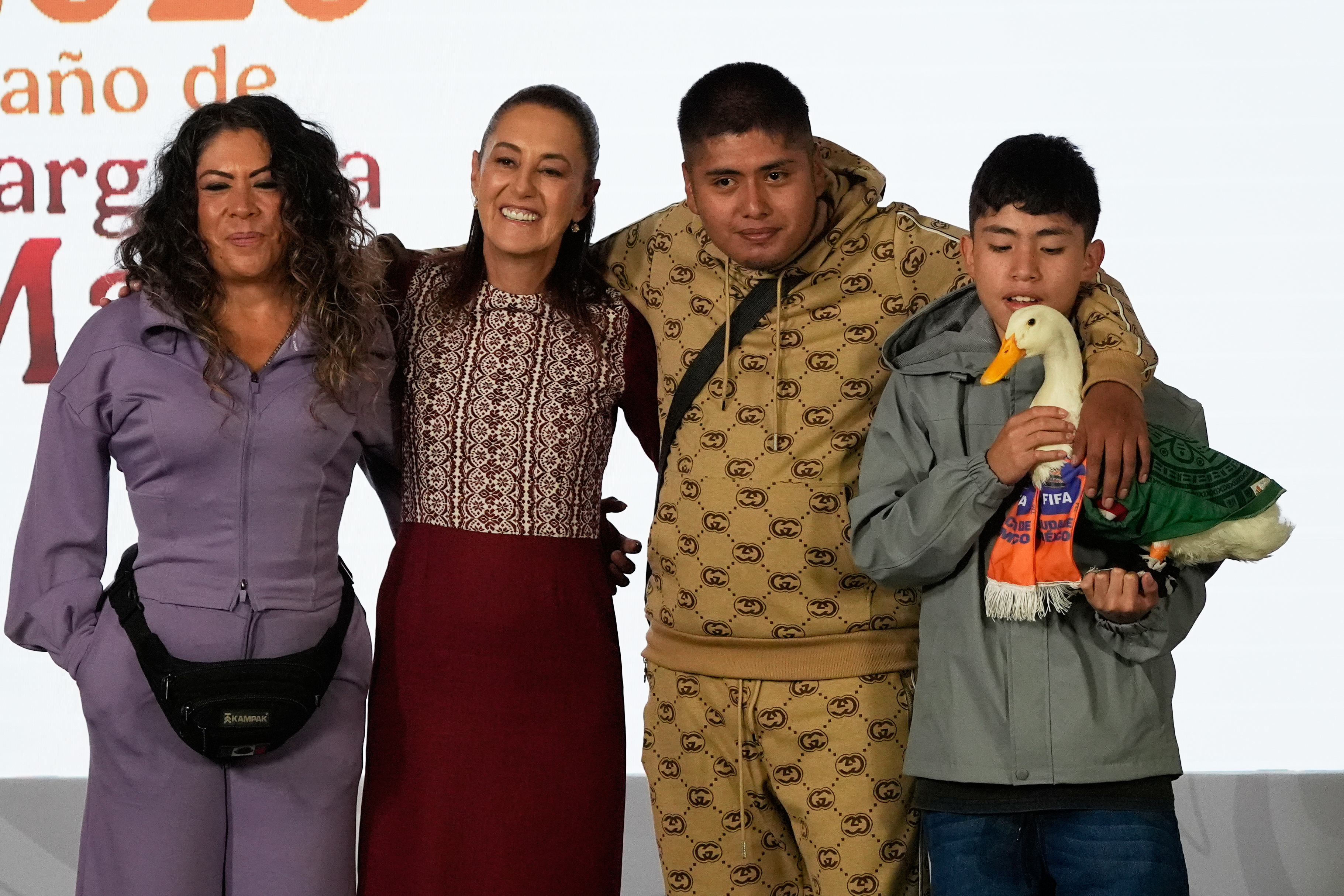 Merlin the duck, dressed in a Mexican national soccer team jersey, attends the daily press conference by Mexican President Claudia Sheinbaum, second from left, along with his caretakers Carla Gomez and her sons Carlos, and Christian who holds Merlin, at the National Palace in Mexico City, Monday, June 22, 2026.