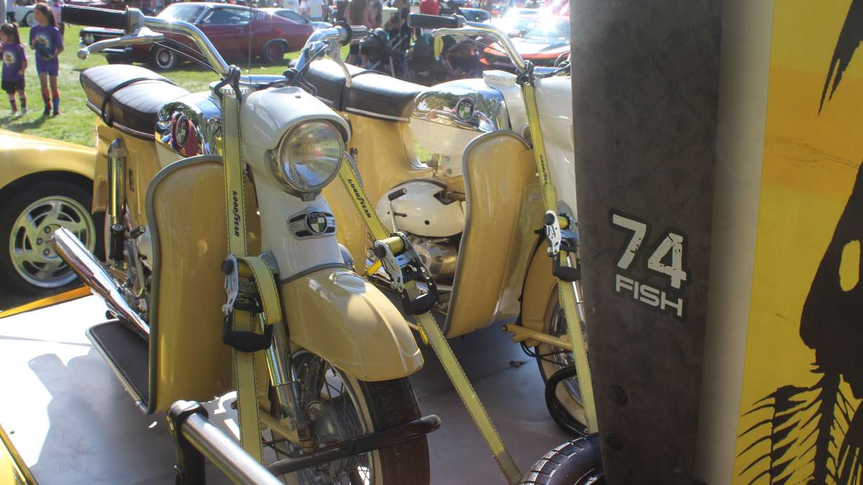 Two 1974 Puch scooters in perfect condition share a trailer with a surfboard that serves as an informational sign describing the scooters and the Jeep pulling them