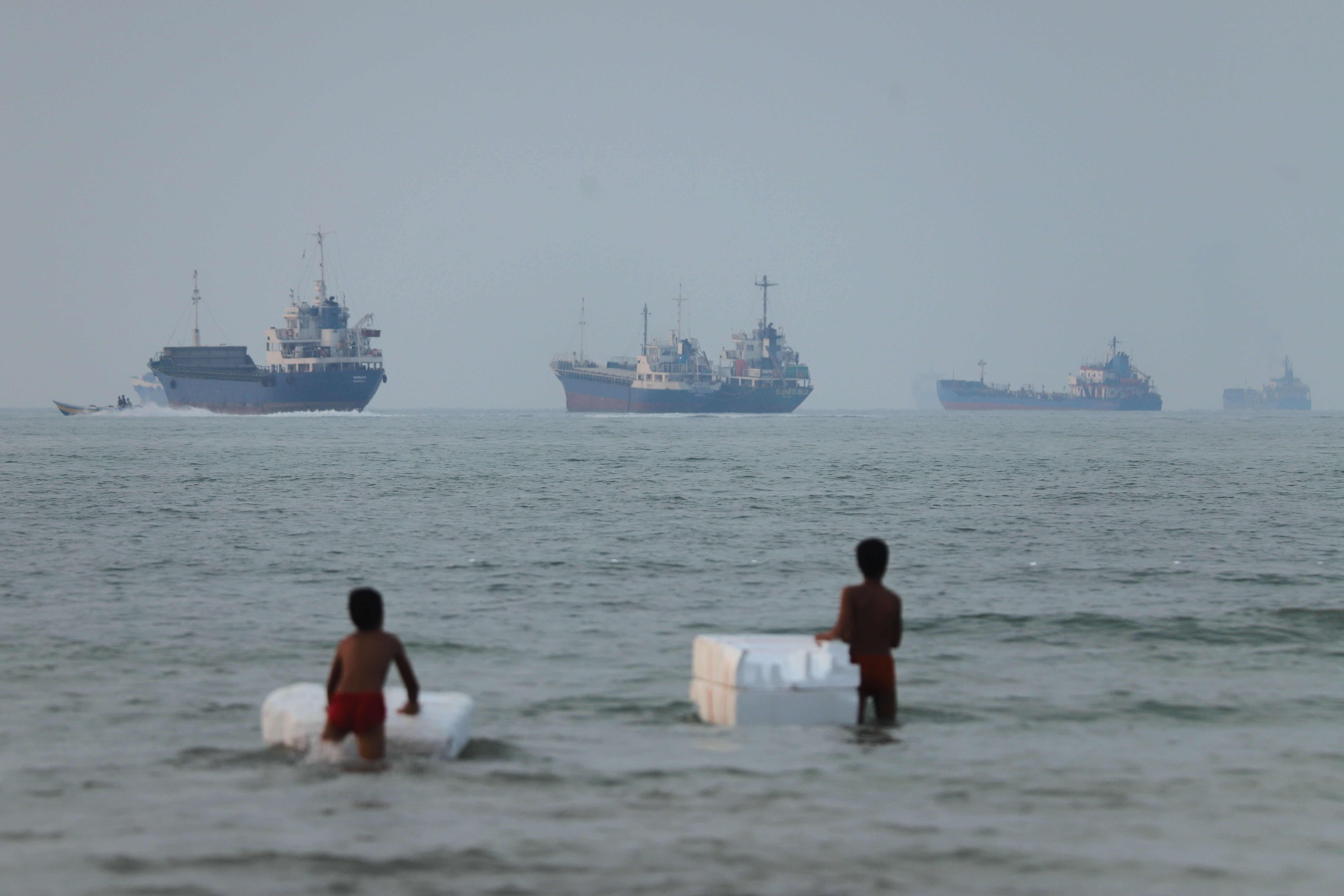 Two boys stand in shallow water with foam floats as cargo ships and other vessels are seen in the Strait of Hormuz off Bandar Abbas, Iran, Tuesday. The strait remains a point of contention as an end to the Iran war is sought.