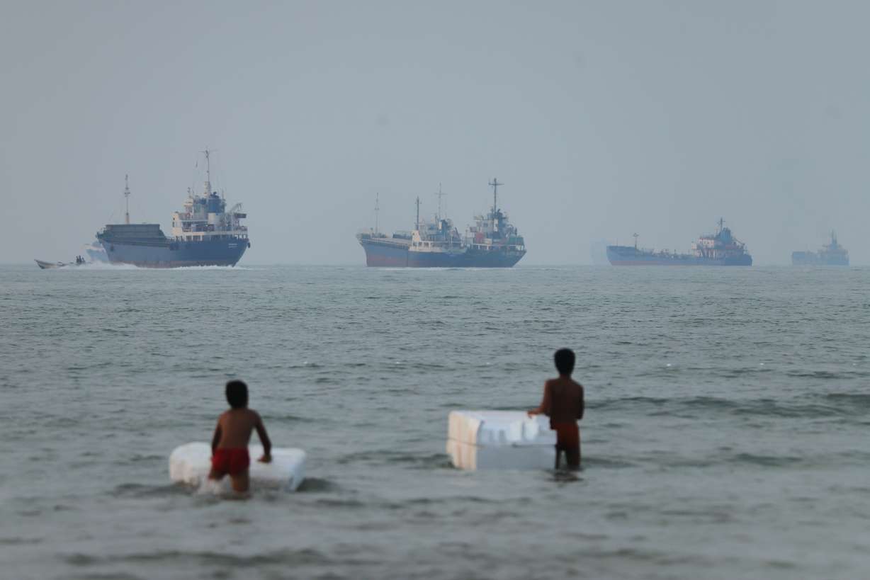 Two boys stand in shallow water with foam floats as cargo ships and other vessels are seen in the Strait of Hormuz off Bandar Abbas, Iran, Tuesday. The strait remains a point of contention as an end to the Iran war is sought.