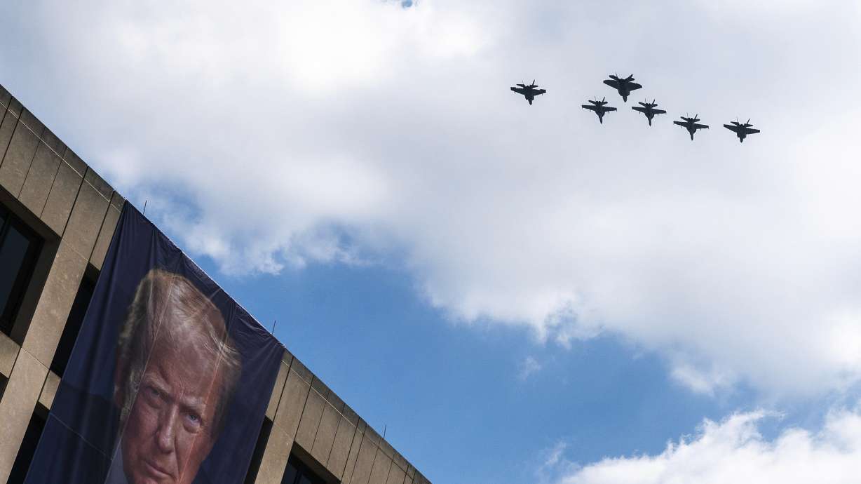 A flight of F-18s, F-35s and a F-22 fly past a picture of President Donald Trump hanging on the Department of Labor near the Great American State Fair on the National Mall, Friday, in Washington. Politics and a heat wave are colliding with Saturday's celebrations.