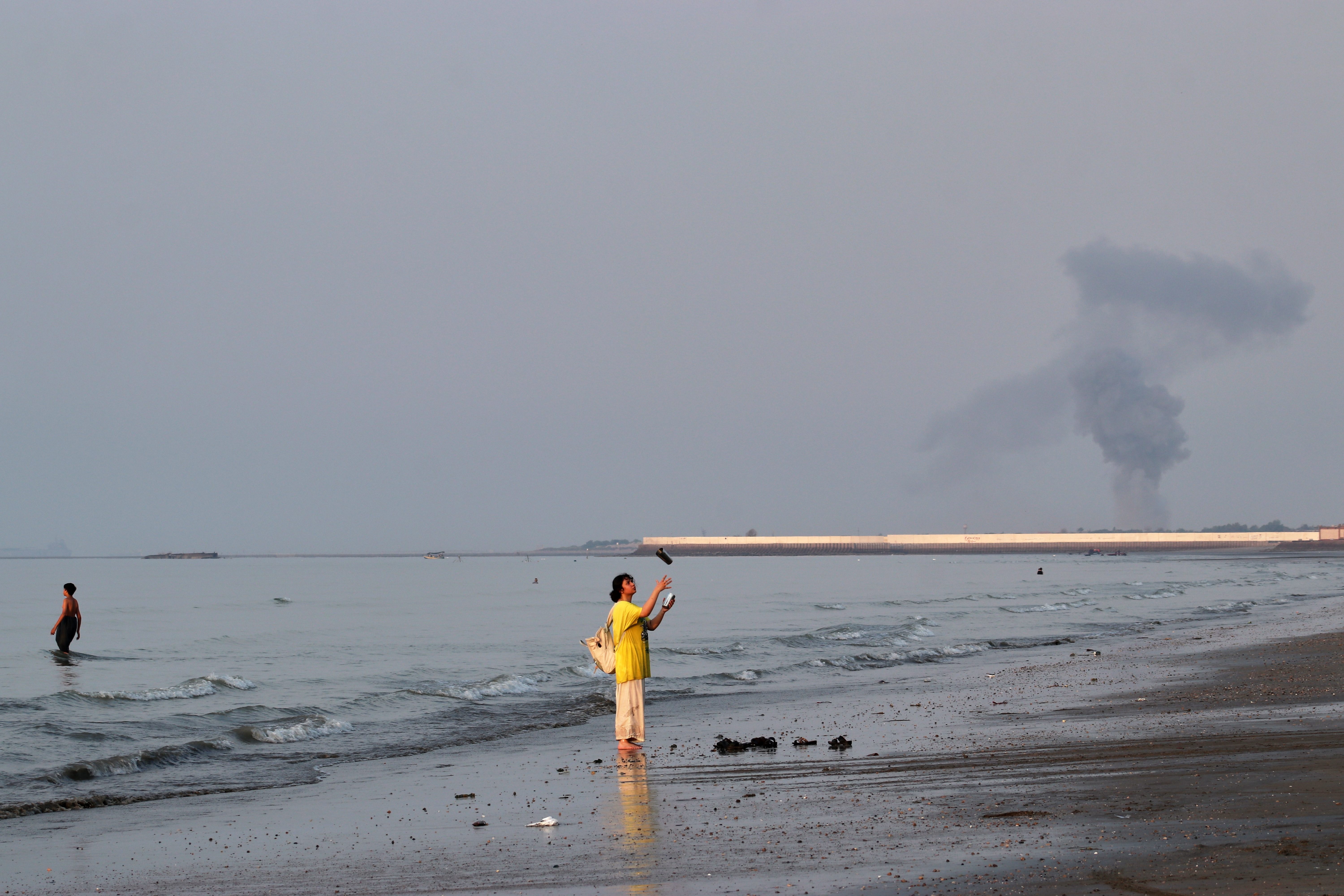 A woman stands at the water's edge along the Strait of Hormuz as a plume of smoke rises in the background following an explosion, off Bandar Abbas, Iran, Monday. The U.S. and Iran continued to trade strikes Thursday as the U.S. expanded its targets northward.