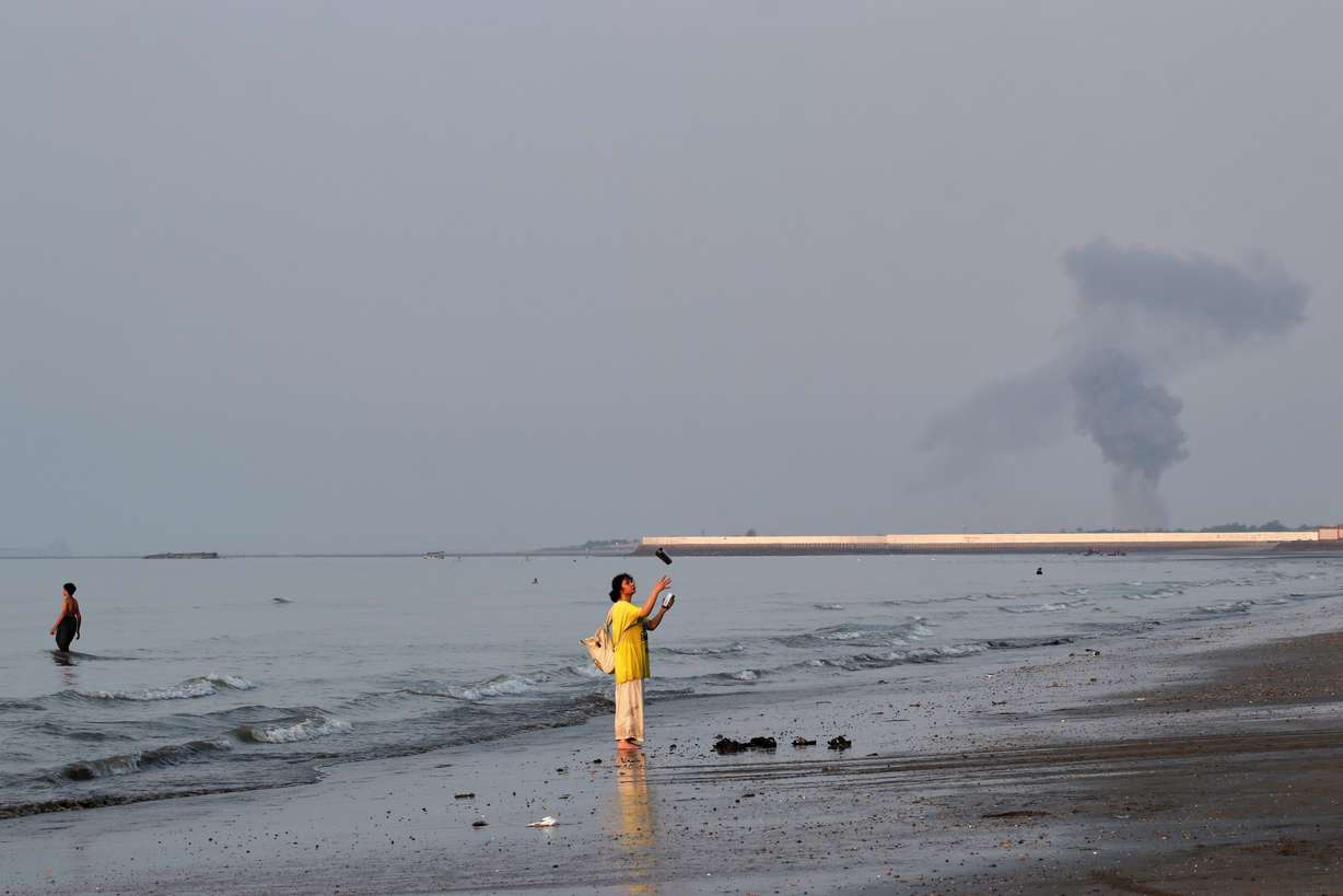 A woman stands at the water's edge along the Strait of Hormuz as a plume of smoke rises in the background following an explosion, off Bandar Abbas, Iran, Monday. The U.S. and Iran continued to trade strikes Thursday as the U.S. expanded its targets northward.