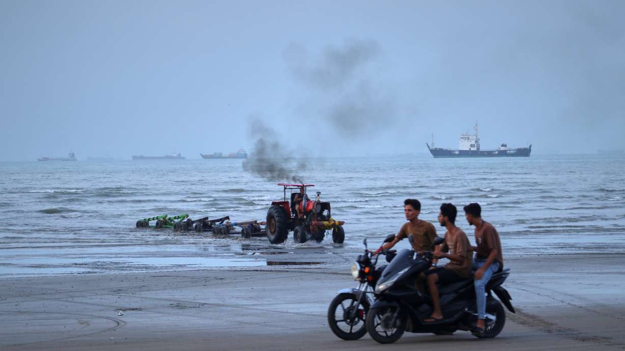 Young men ride motorcycles along the shore as a tractor pulls equipment from the water and commercial vessels appear anchored in the Strait of Hormuz off Bandar Abbas, Iran, Monday.