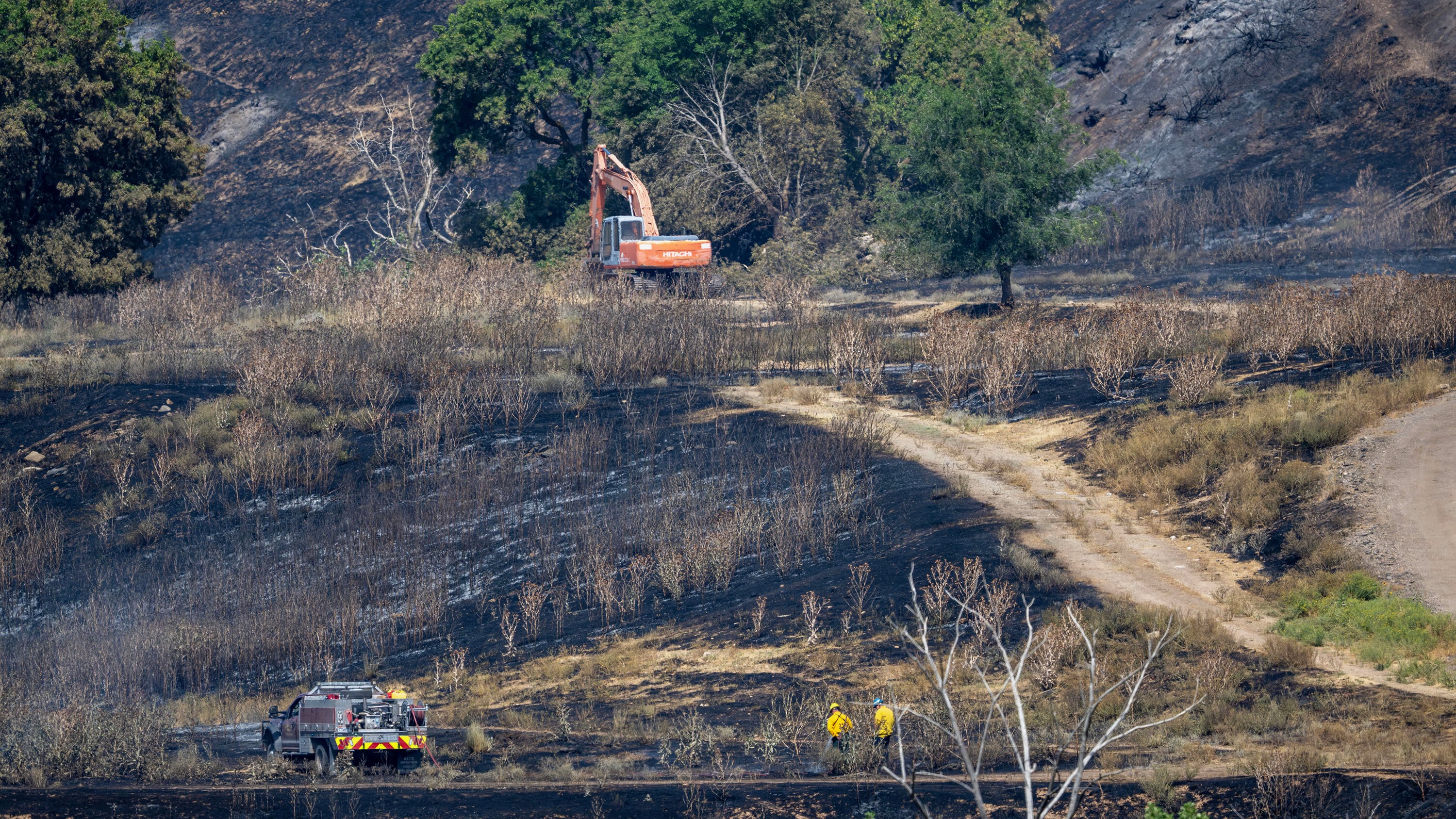 An excavator moves debris as firefighters keep watch on Tuesday of the area torched in a fire Monday in Washington Terrace.
