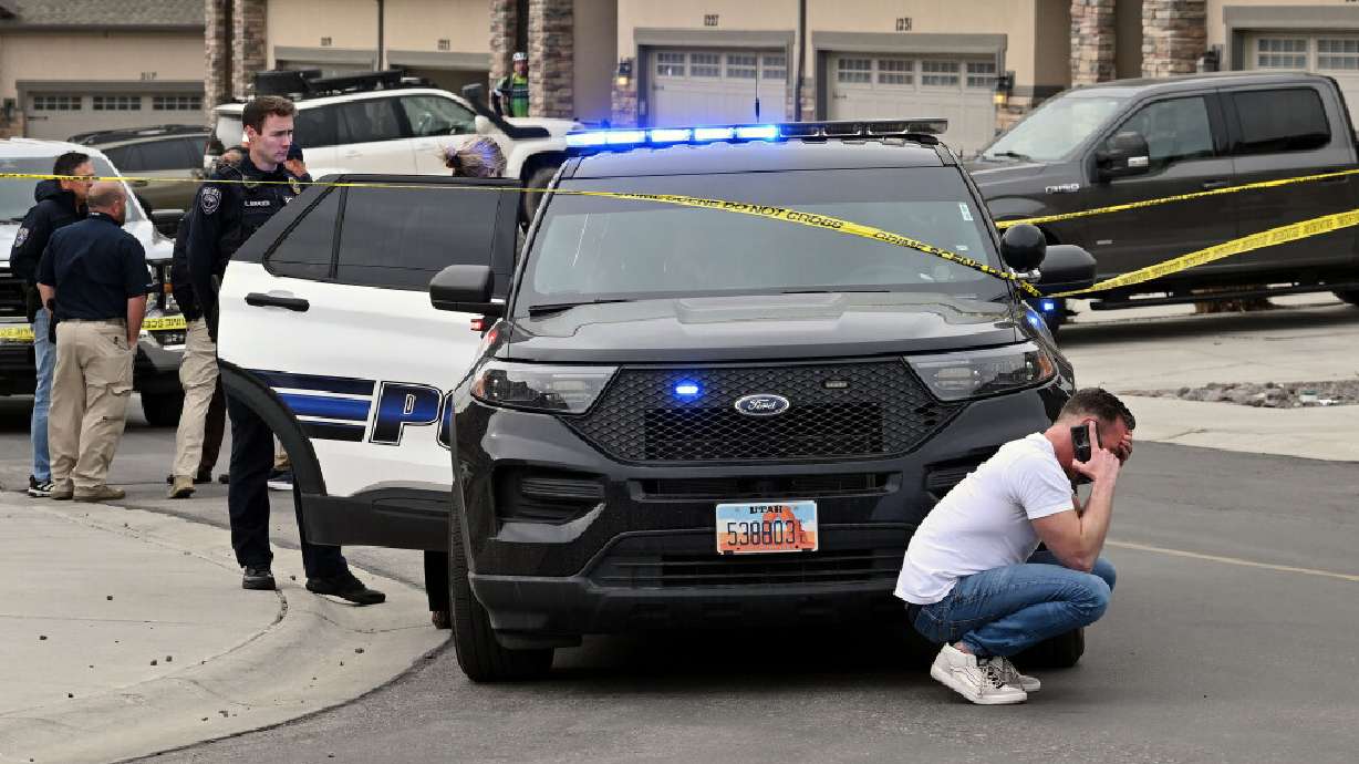 An unidentified man crouches down in front of a police vehicle talking on a cellphone as Saratoga Springs Police investigate deaths of two people in a home on March 28, 2025.