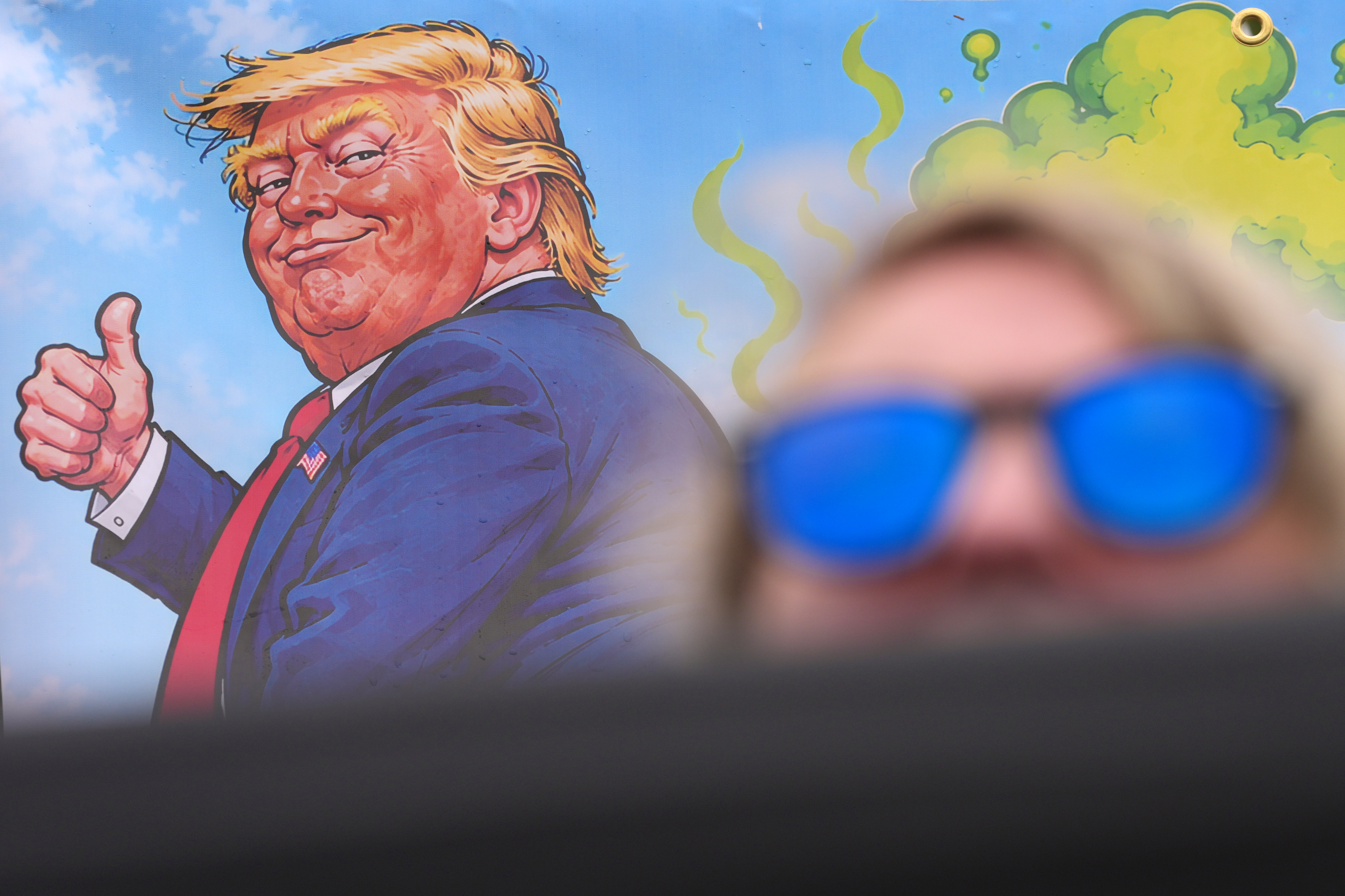 A protester wears sunglasses and sits in front of a poster depicting U.S. President Donald Trump during his visit to the Irish Open at the Trump International Golf Links course in Doonbeg, Ireland, Sunday, Sept. 13, 2026.