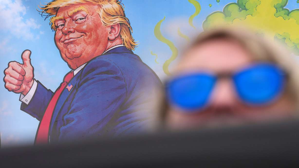 A protester wears sunglasses and sits in front of a poster depicting U.S. President Donald Trump during his visit to the Irish Open at the Trump International Golf Links course in Doonbeg, Ireland, Sunday, Sept. 13, 2026.