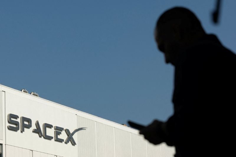 SpaceX logo as an employe looks at his phone while making his way to work at the company’s facility on the day of the SpaceX IPO, in Hawthorne, California, U.S. June 12, 2026.