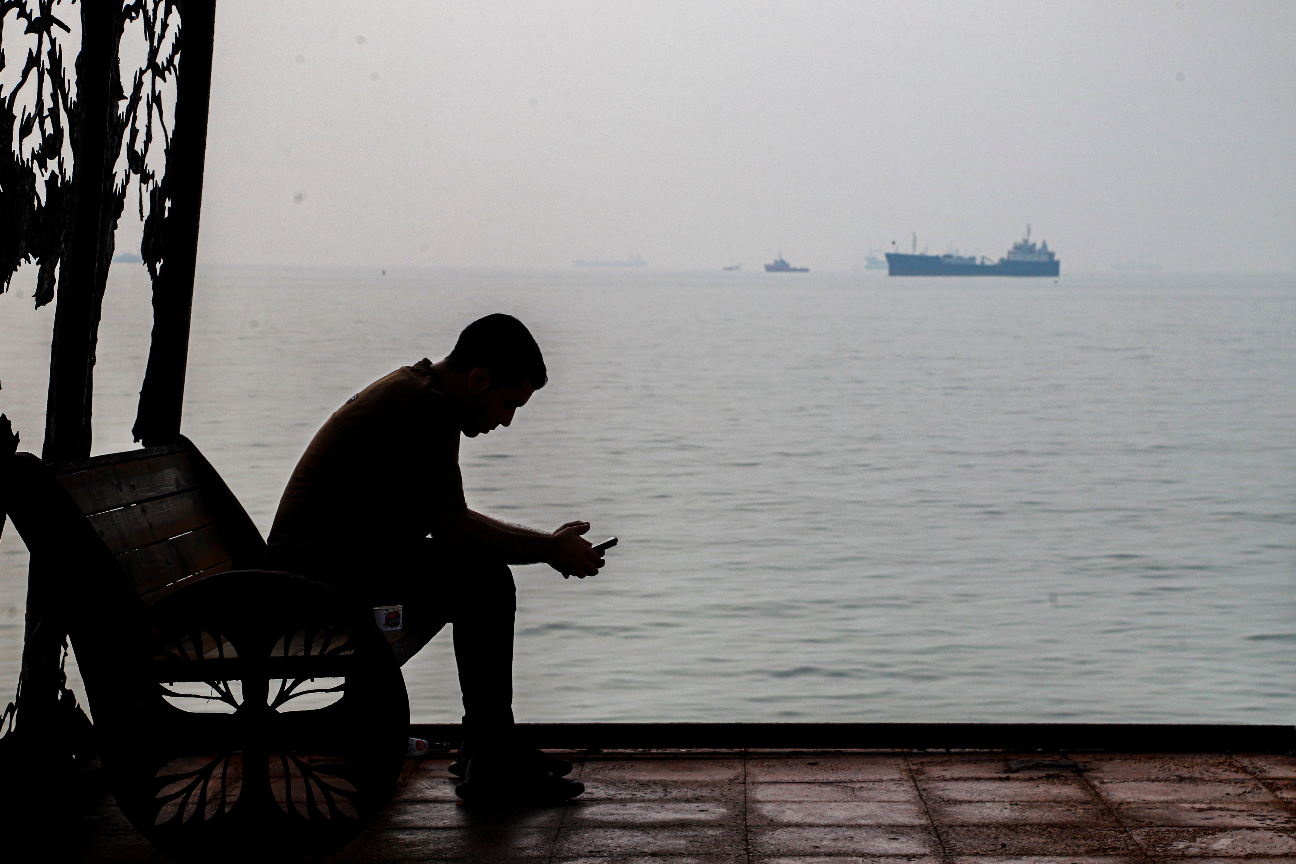 A man checks his cellphone at a waterfront shelter as commercial vessels appear anchored in the Strait of Hormuz off Bandar Abbas, Iran, Oct. 6. A think tank said on Tuesday that Iran may be targeting ship-to-ship transfers in the strait.