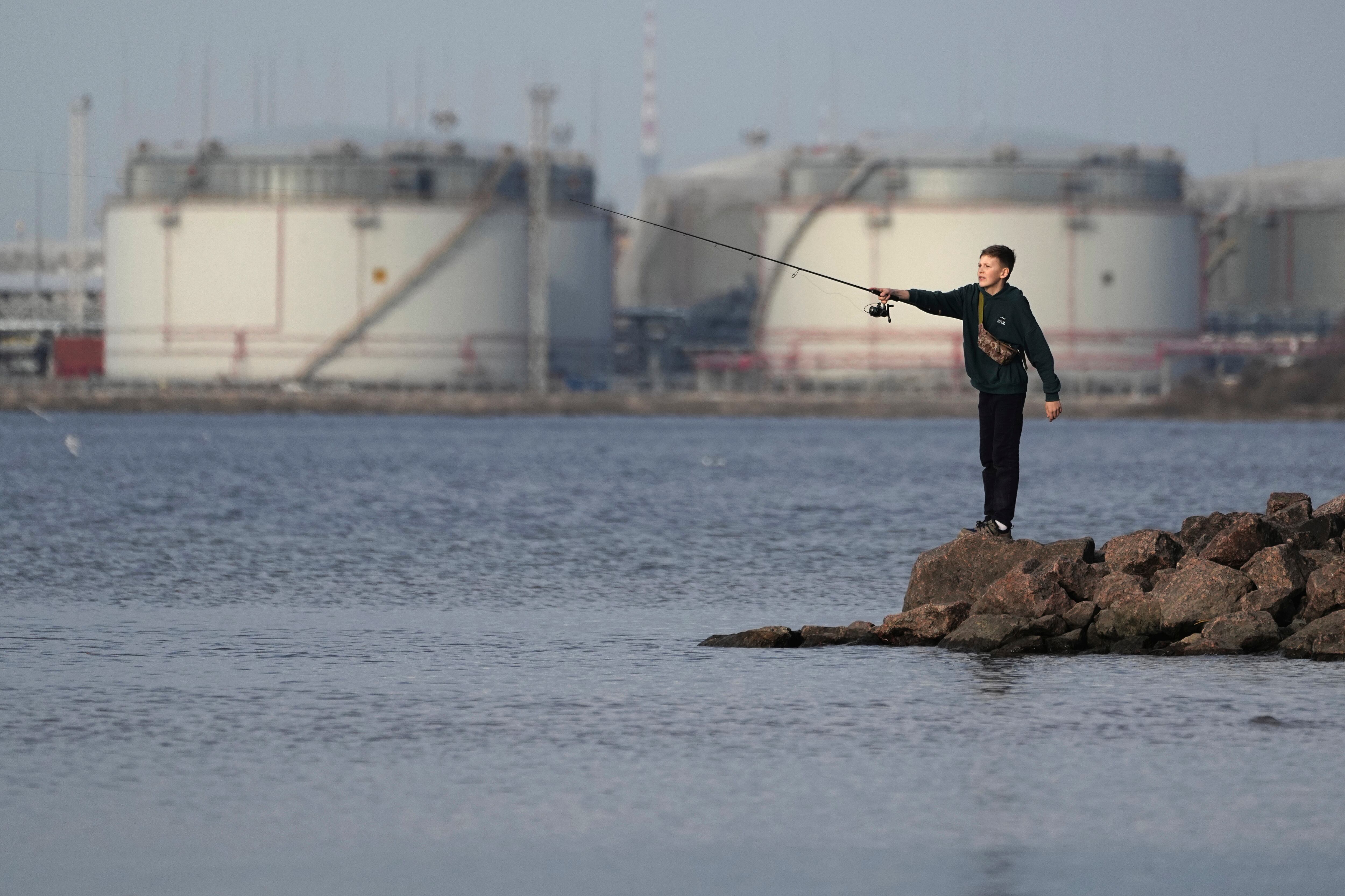 A boy fishes on the coast of the Gulf of Finland in St. Petersburg, Russia, Sunday, March 30, 2025, with oil storage tanks in the port of St. Petersburg in the background.